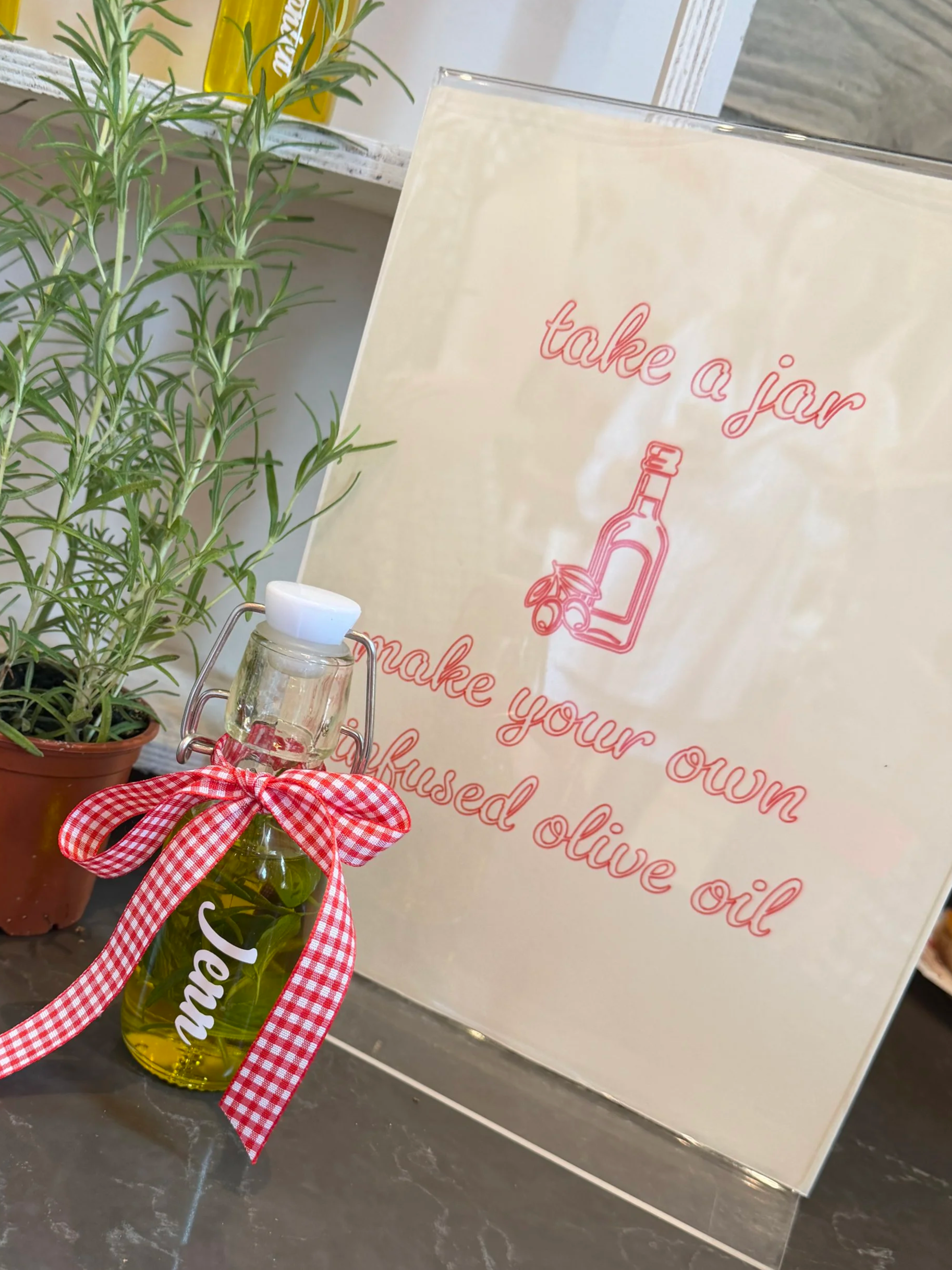 A bottle of olive oil with a red and white checkered ribbon tied around its neck, sitting on a black marble surface next to a potted herb, with a sign in the background that says 'take a jar' and promotes olive oil.
