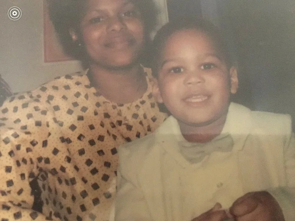 A photograph of small, Black male child dressed in a white suit with an adult, Black woman in a checkered top smiling