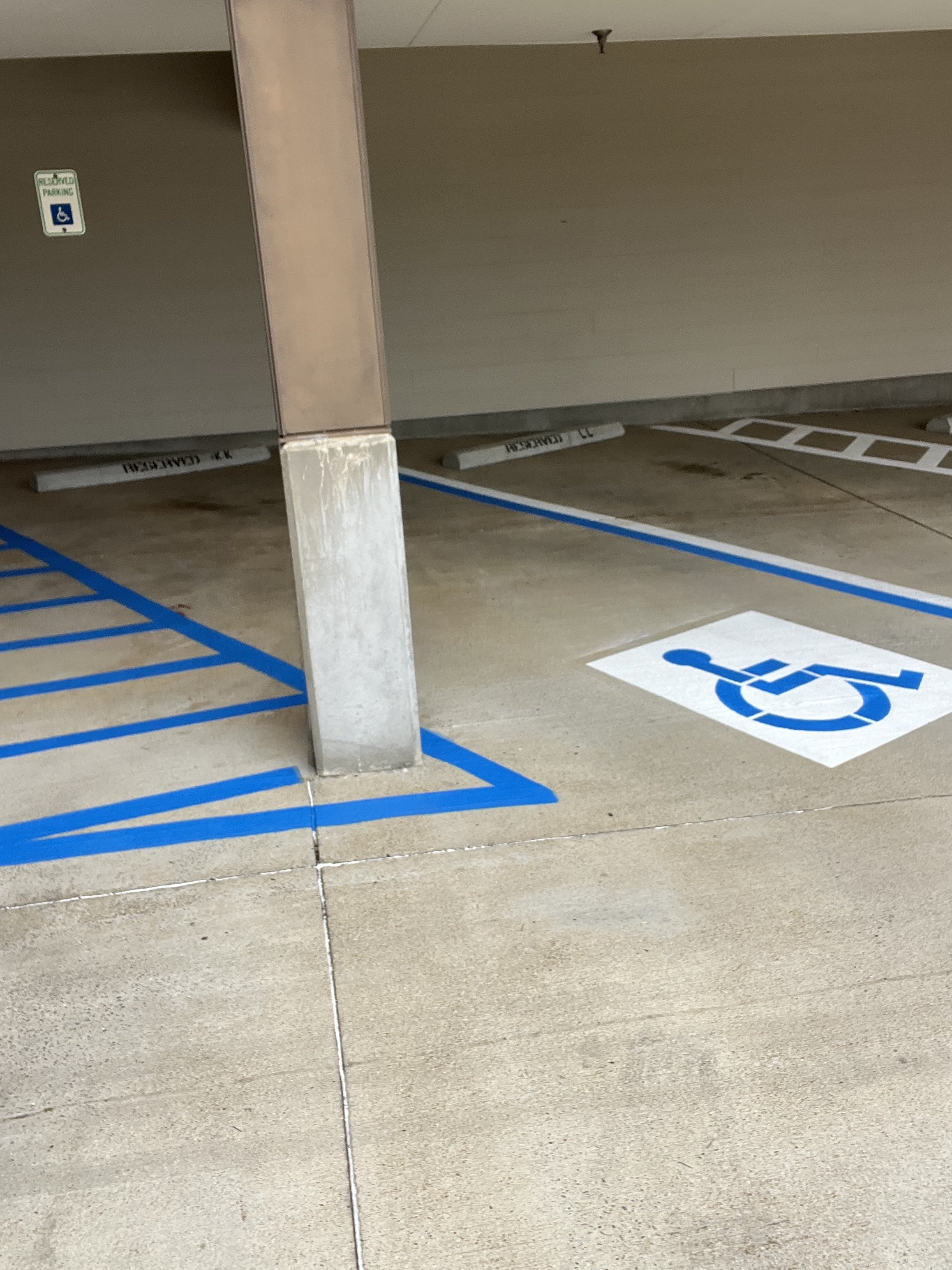 An outdoor parking space with designated handicapped parking spots marked by blue paint and a wheelchair symbol on the ground. There's a concrete pole and a wall with a sign indicating accessible parking.
