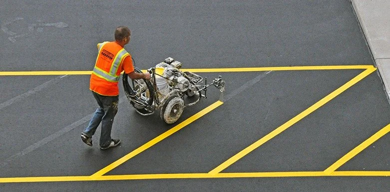 A worker in an orange safety vest operates a line marking machine to paint yellow lines on a dark asphalt surface, creating parking lot markings.