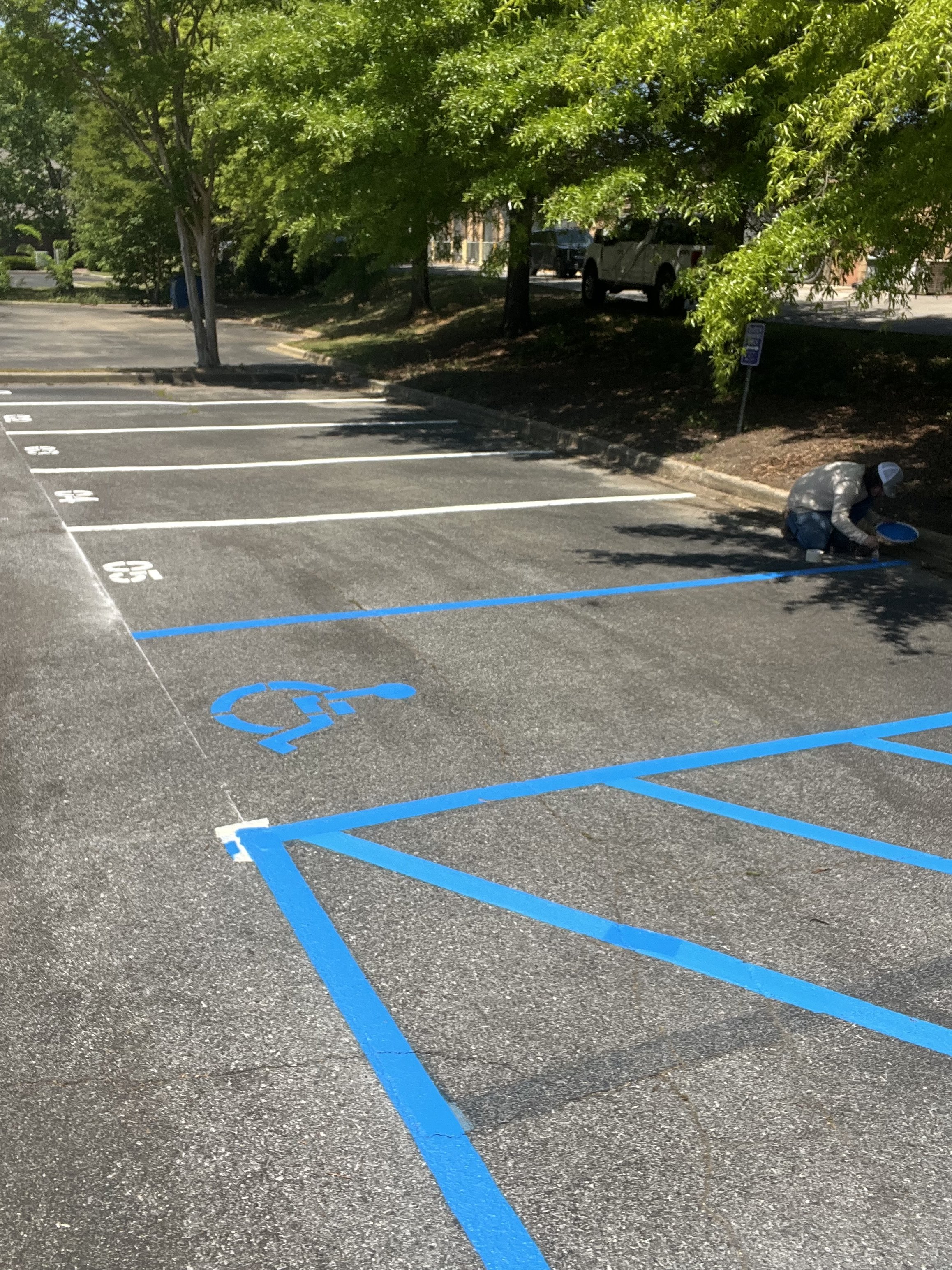 A person in a cap and jacket is painting blue lines to designate a handicapped parking space in a parking lot, with green trees and a parked truck in the background.