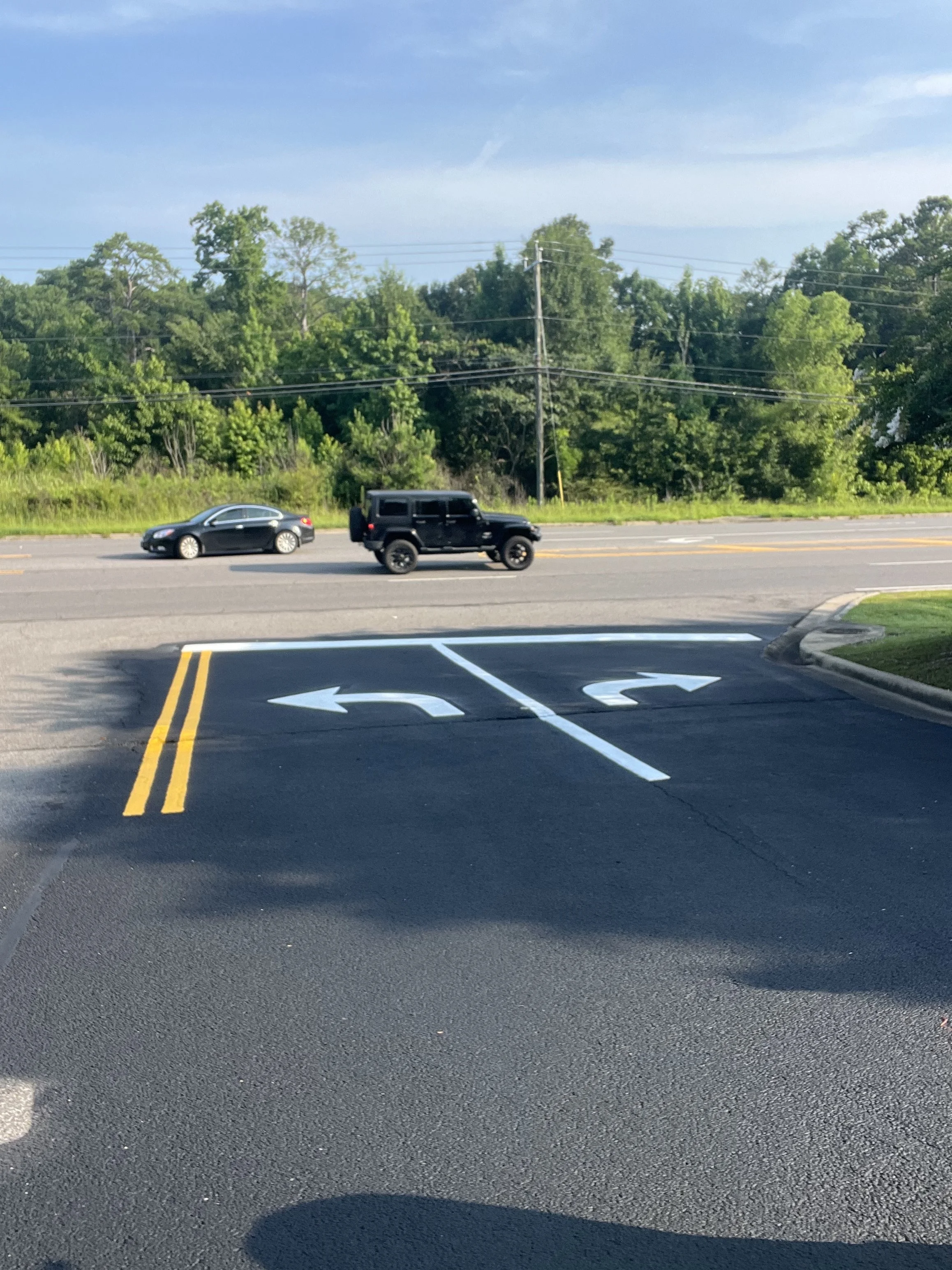 Parking lot view showing two cars on a road, with a double yellow line, and directional arrows painted on the asphalt, and a green treeline in the background under a blue sky.