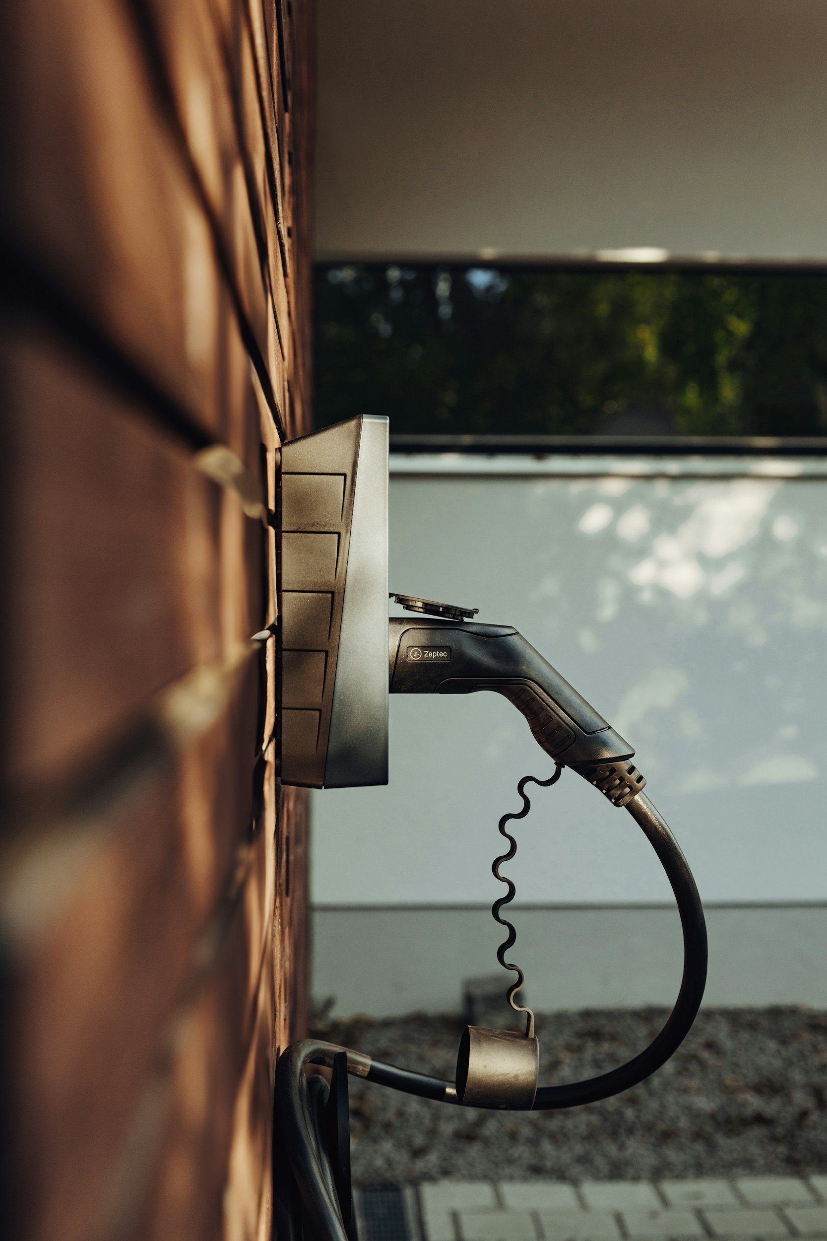 Close-up of an electric curling iron plugged into an outlet on a wooden wall, with a window showing trees outside in the background.
