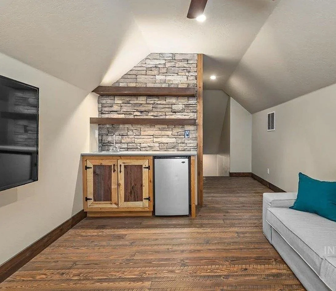 A small living area with a built-in kitchenette, featuring a stone wall, wooden shelves, a mini fridge, and a sink. To the right is a white sofa with a blue pillow, and on the left side is a mounted flat-screen TV. The space has wood flooring and slo