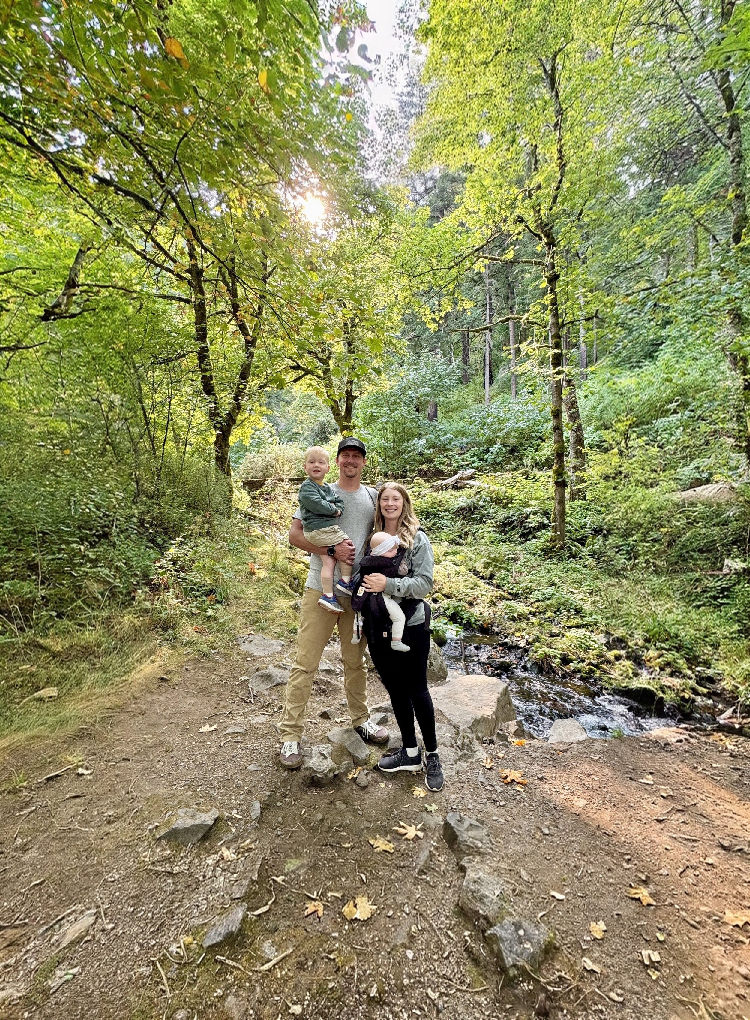 A family of four, including a man, a woman, a boy, and a baby, posing on a trail in a lush, green forest near a small creek.