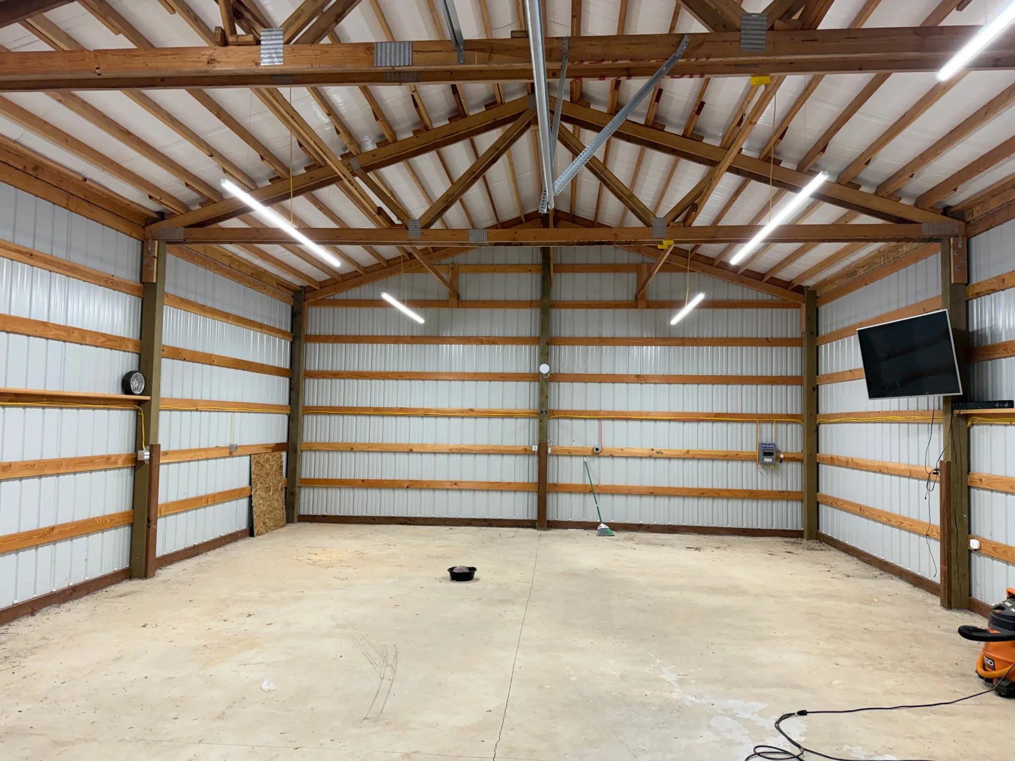 Interior of a large unfinished building with exposed wooden beams, metal siding walls, concrete floor, and modern fluorescent lighting. There is a black bucket on the floor and some tools visible.