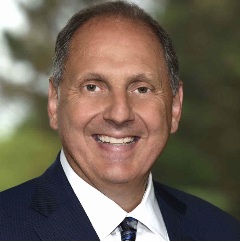 A middle-aged man with short brown hair, smiling, dressed in a navy suit, white shirt, and striped tie, standing outdoors with a blurred green background.