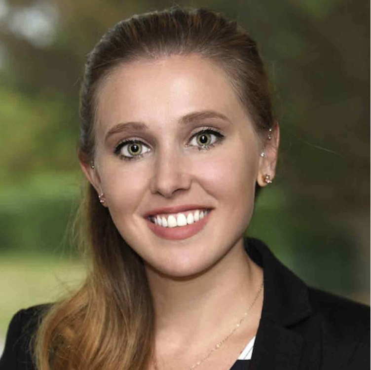 A smiling woman with long, light brown hair, green eyes, and earrings, outdoors with a blurred green background.
