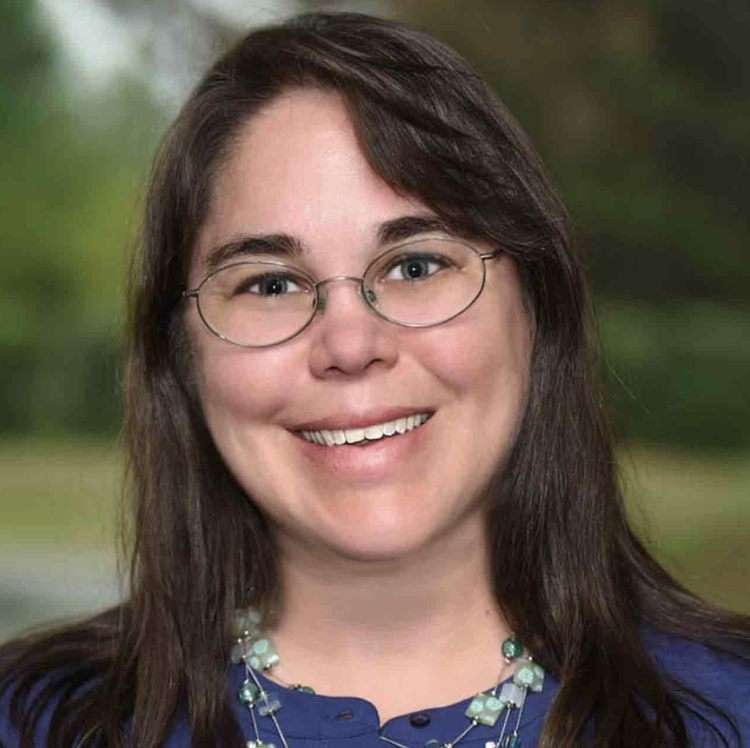 A woman with long brown hair, glasses, and a blue top, smiling outdoors with a blurred green background.