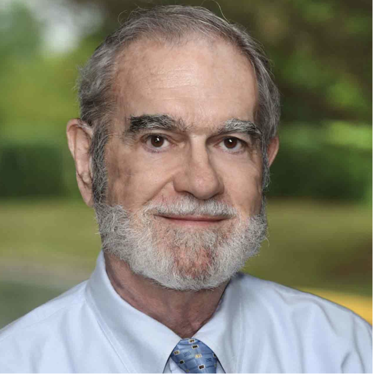 Close-up portrait of an older man with gray hair and beard, wearing a light blue dress shirt and a patterned tie, standing outdoors with a blurred green background.