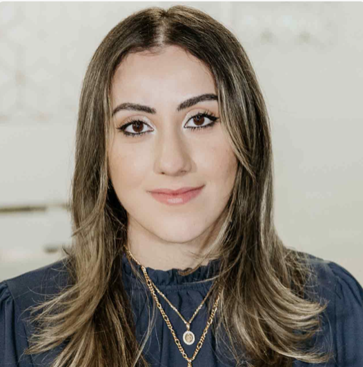 Young woman with shoulder-length brown hair, light makeup, wearing a navy blouse and layered necklaces, standing against a light-colored background.