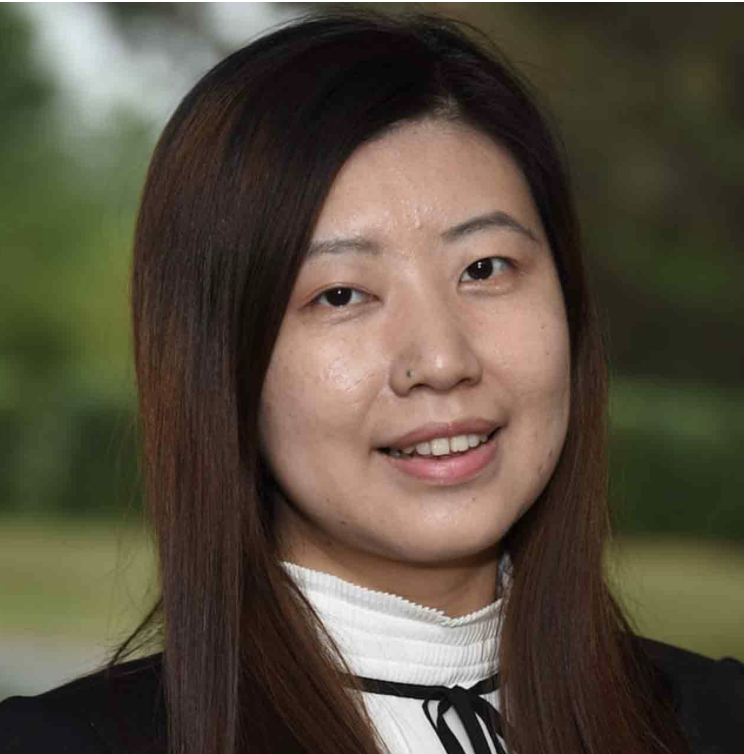 A young woman with straight, dark brown hair smiling outdoors, wearing a white turtleneck and black top.