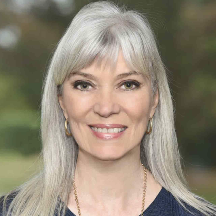 A woman with long gray hair, wearing gold hoop earrings and a gold necklace, smiling outdoors with a blurred green background.