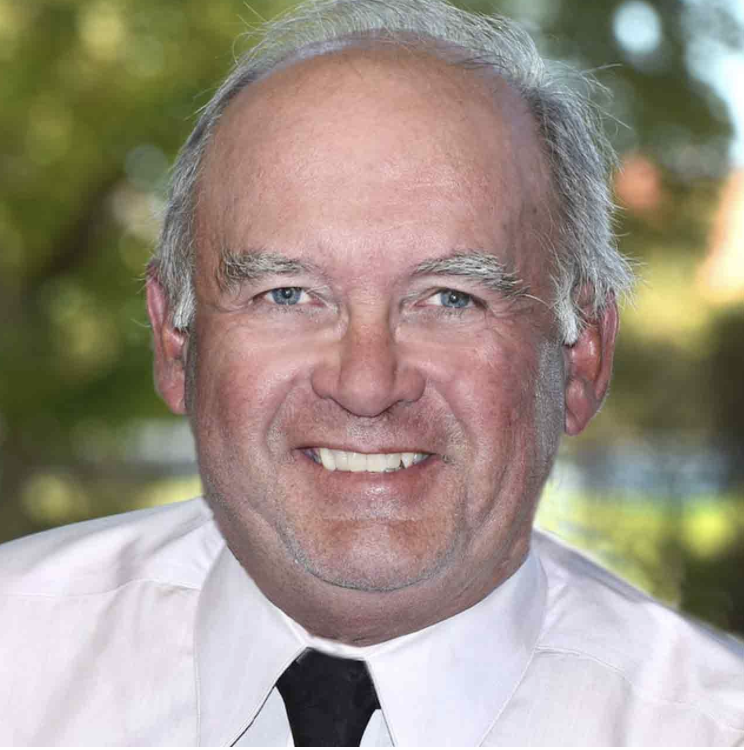 Smiling middle-aged man with gray hair and blue eyes wearing a white shirt and black tie, outdoors with blurred greenery background.