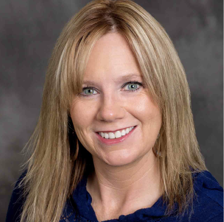 Headshot of a smiling woman with blonde hair and blue eyes, wearing a dark blue top against a gray background.
