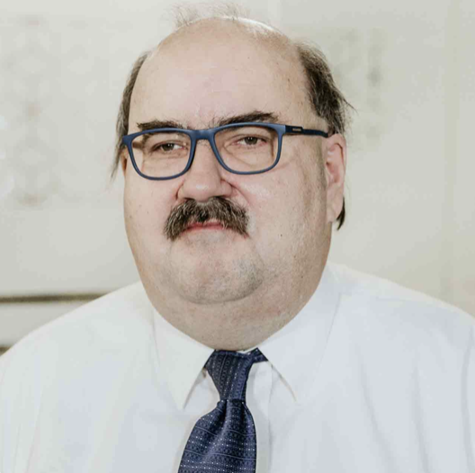 Close-up of a middle-aged man with glasses, a mustache, a white collared shirt, and a navy blue tie, in an indoor setting.