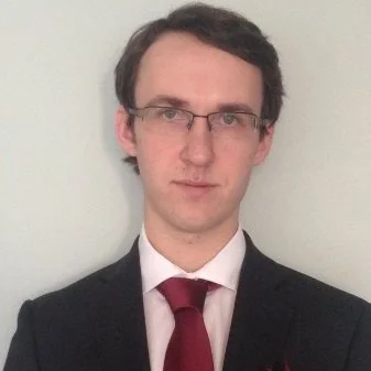 A young man wearing glasses, a dark suit, white shirt, and red tie standing against a plain white wall.