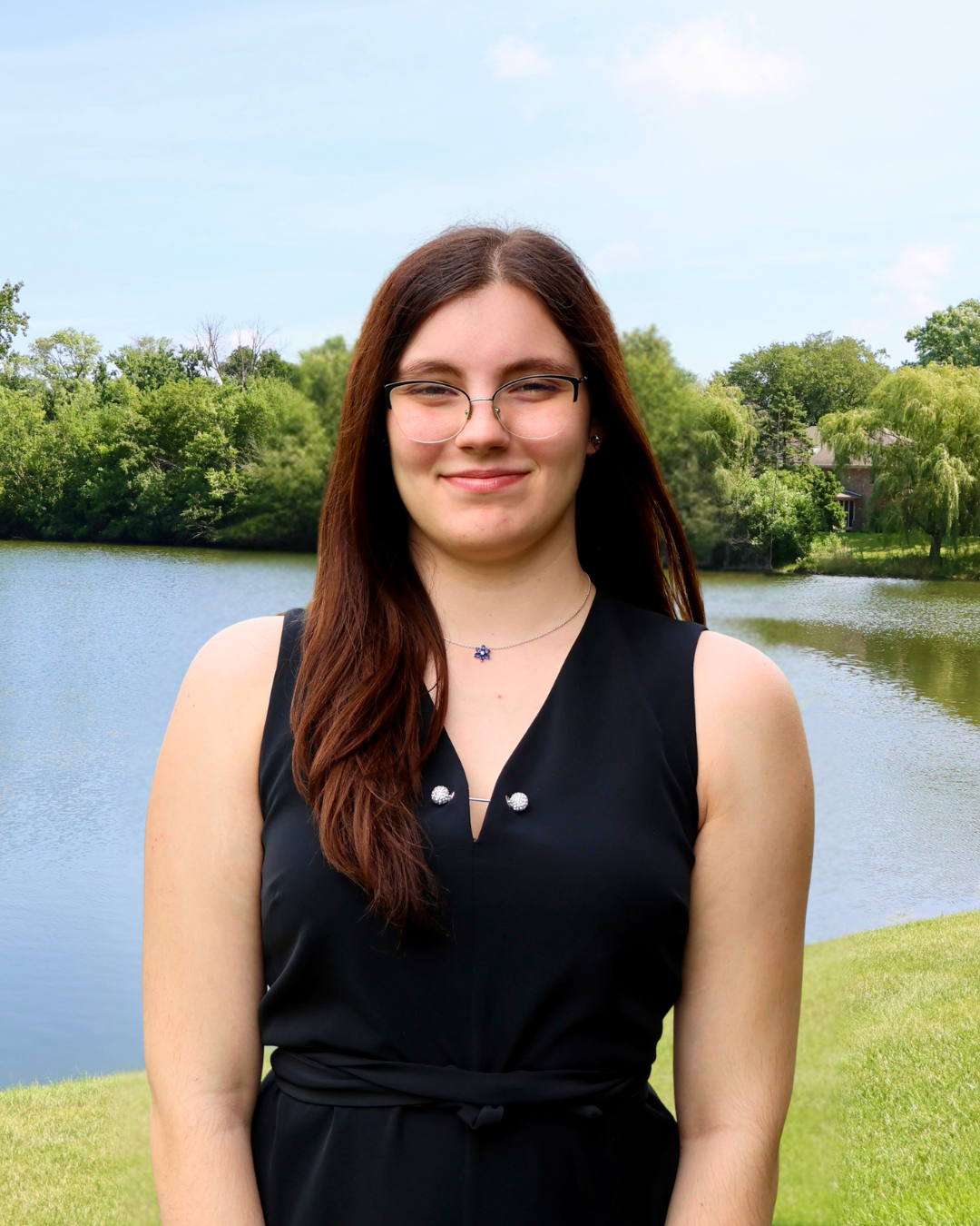 Young woman with brown hair, glasses, and a black sleeveless dress, standing outdoors next to a body of water with trees and houses in the background on a partly cloudy day.