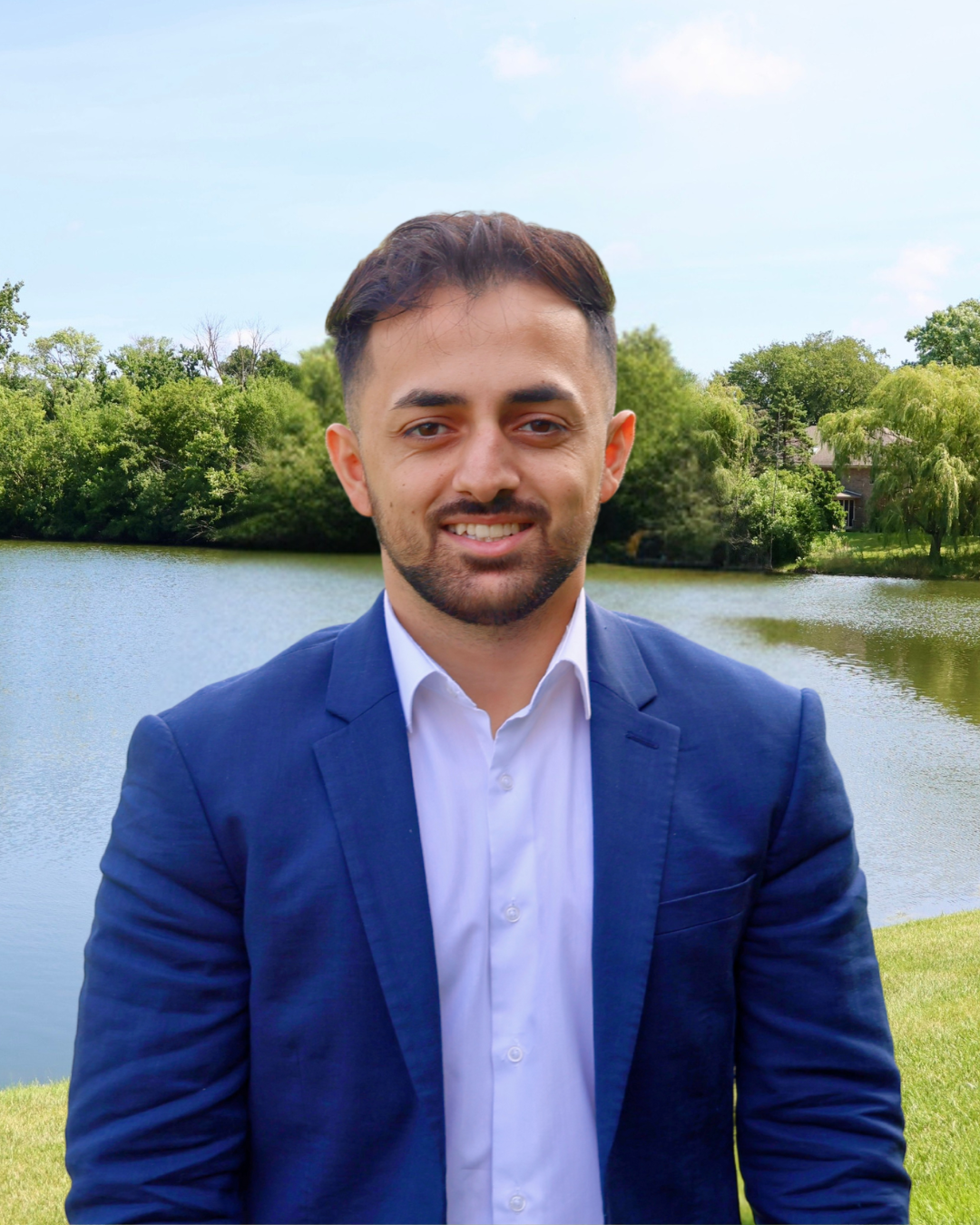 A man in a blue suit and white shirt standing outdoors near a lake with green trees and a house in the background.