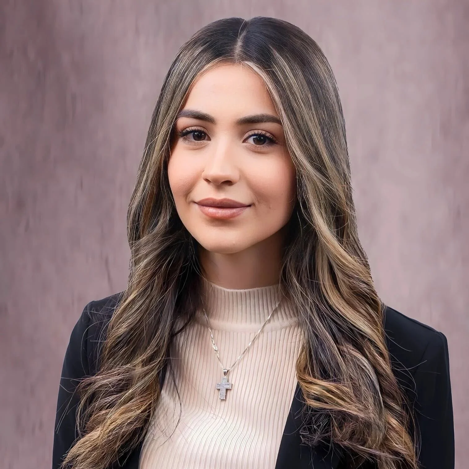 Portrait of a young woman with long, wavy brown hair with blonde highlights, wearing a black blazer over a beige top, and a silver cross necklace. She has a slight smile and makeup, standing against a muted pink background.
