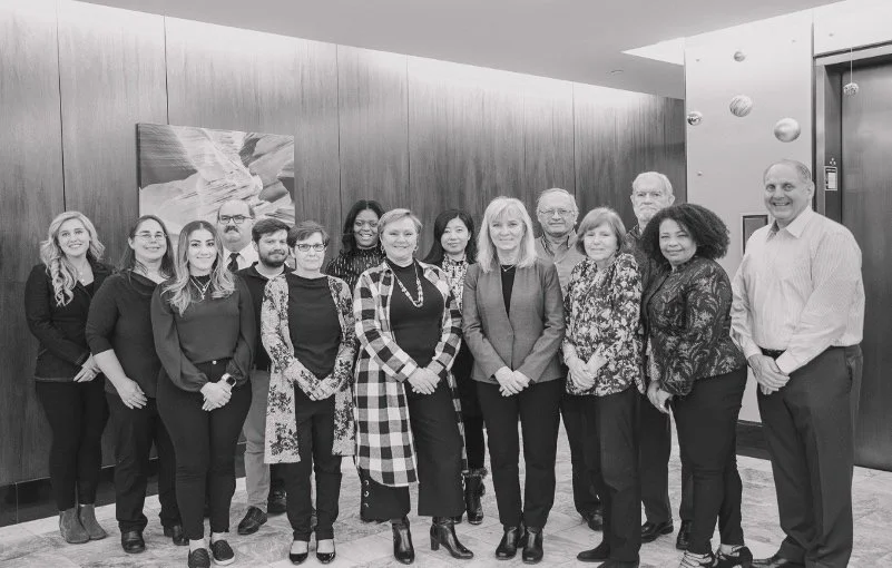 Group of 14 diverse professionals, male and female, standing together in an indoor setting with wood-paneled walls and modern decor, posing for a photo.
