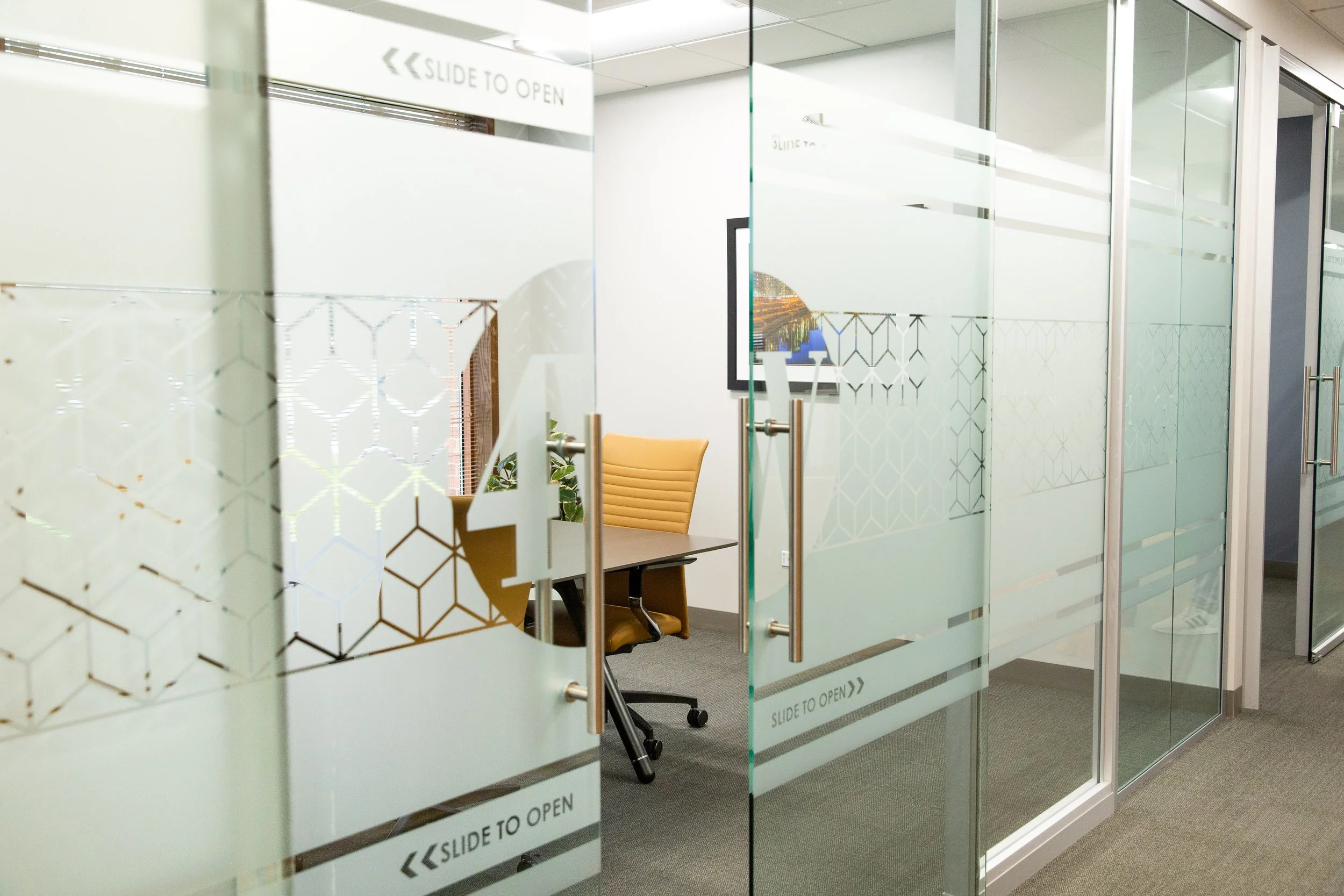 Interior of an office with frosted glass walls and a door labeled 'Slide to Open,' a framed picture on the wall, a yellow office chair, and potted plants.