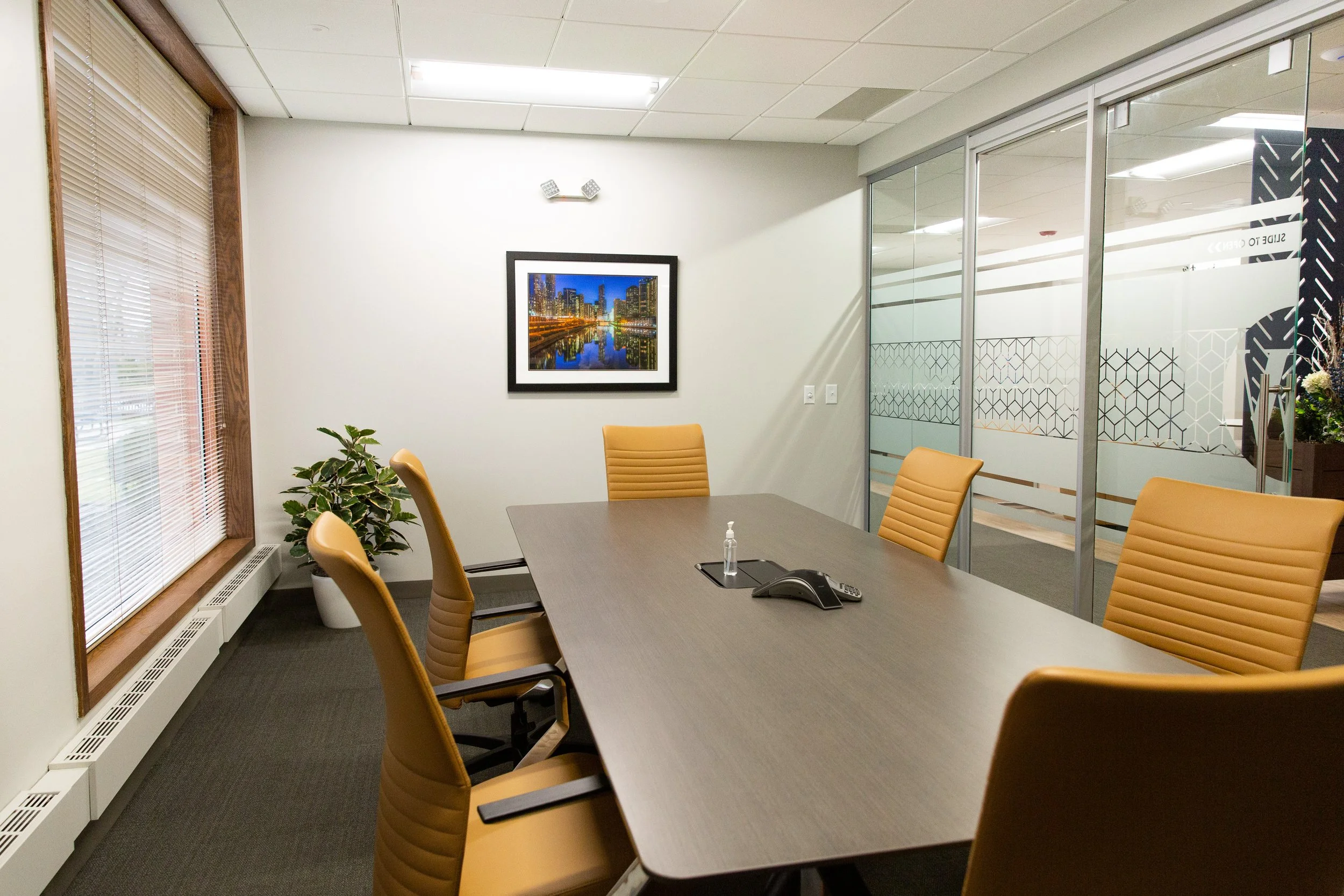 A conference room with beige chairs around a large table, a plant in a white pot, a framed cityscape picture on the white wall, and glass partitions with geometric patterns.