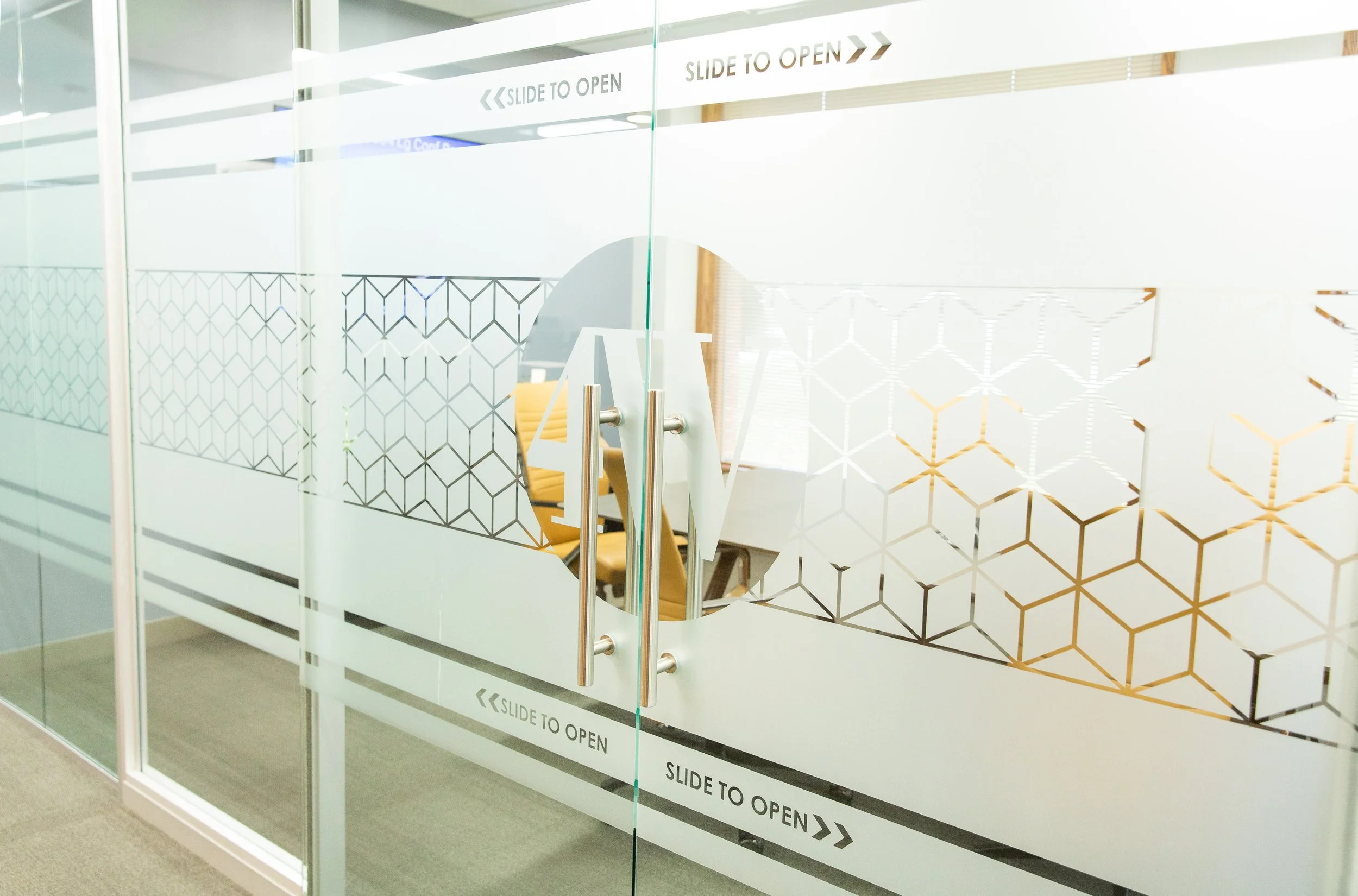 Glass door with black geometric pattern and white sliding door handle, with signage suggesting it slides to open. Inside the room, yellow and brown chairs are visible.