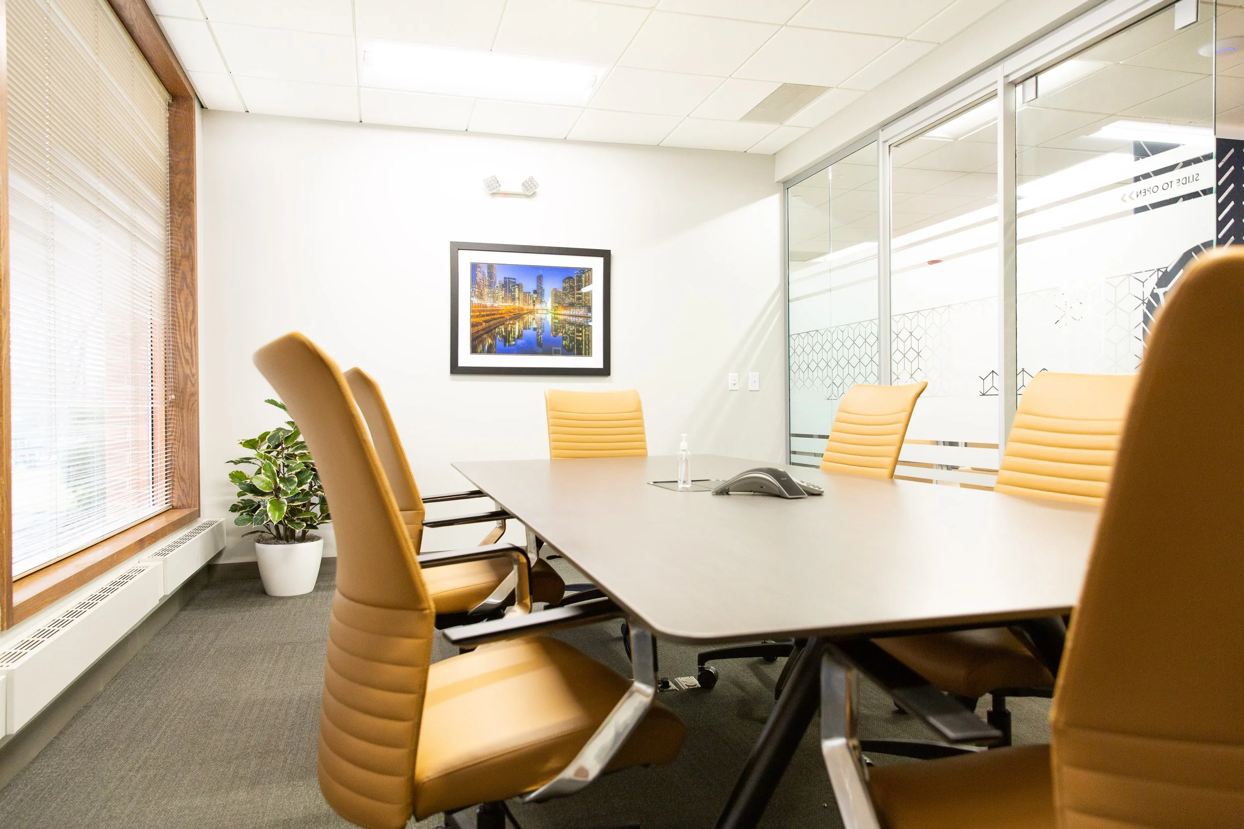 Empty conference room with beige chairs around a table, a wall-mounted picture of a city skyline at night, a potted plant, and large windows with blinds.