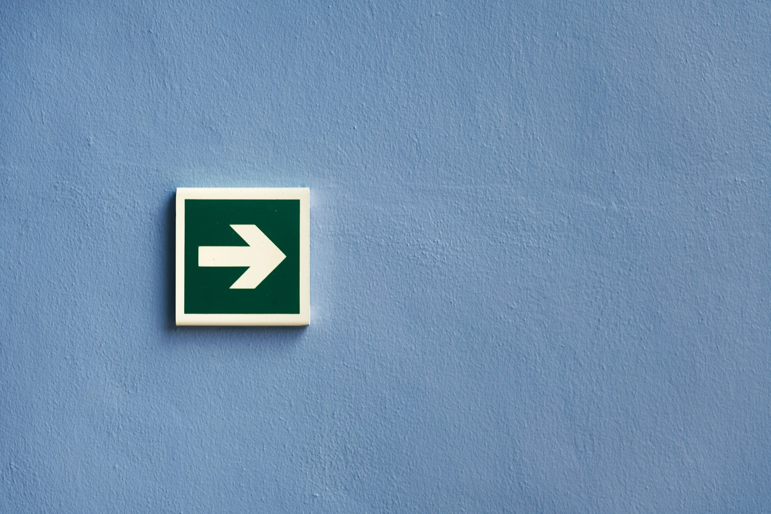 Green emergency exit sign with a white right arrow mounted on a light blue textured wall.