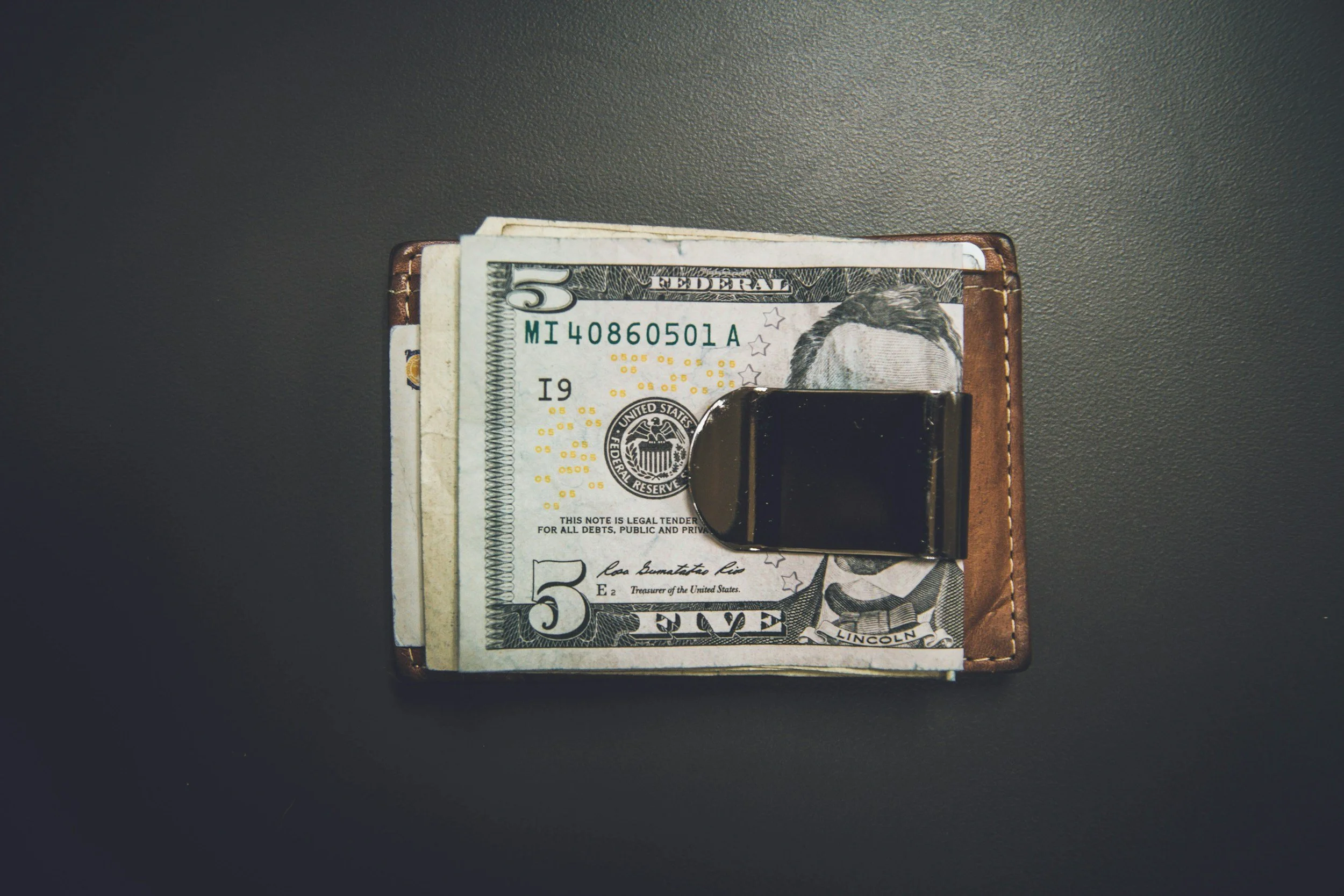 A brown leather wallet with United States five-dollar bills and a black clip inside, placed on a dark gray surface.