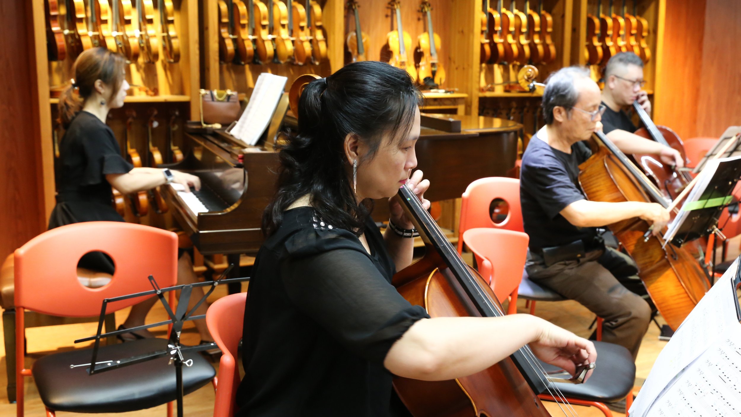 A group of four musicians playing string instruments, including a woman playing a cello, in a music rehearsal room with violins hanging on the wooden wall behind them.