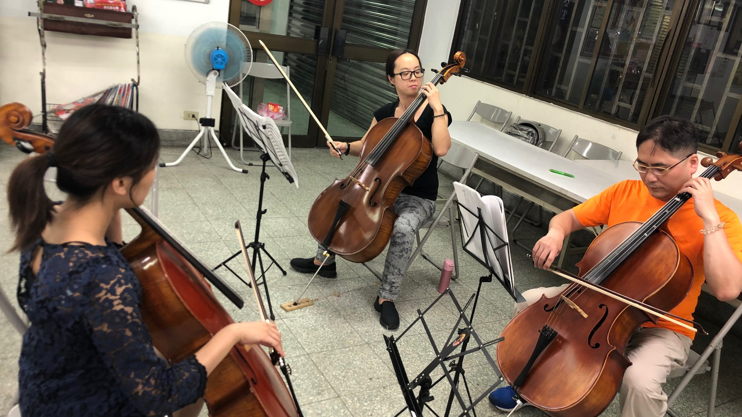 Four musicians playing string instruments, two women and two men, in an indoor rehearsal space with music stands, chairs, and a fan.