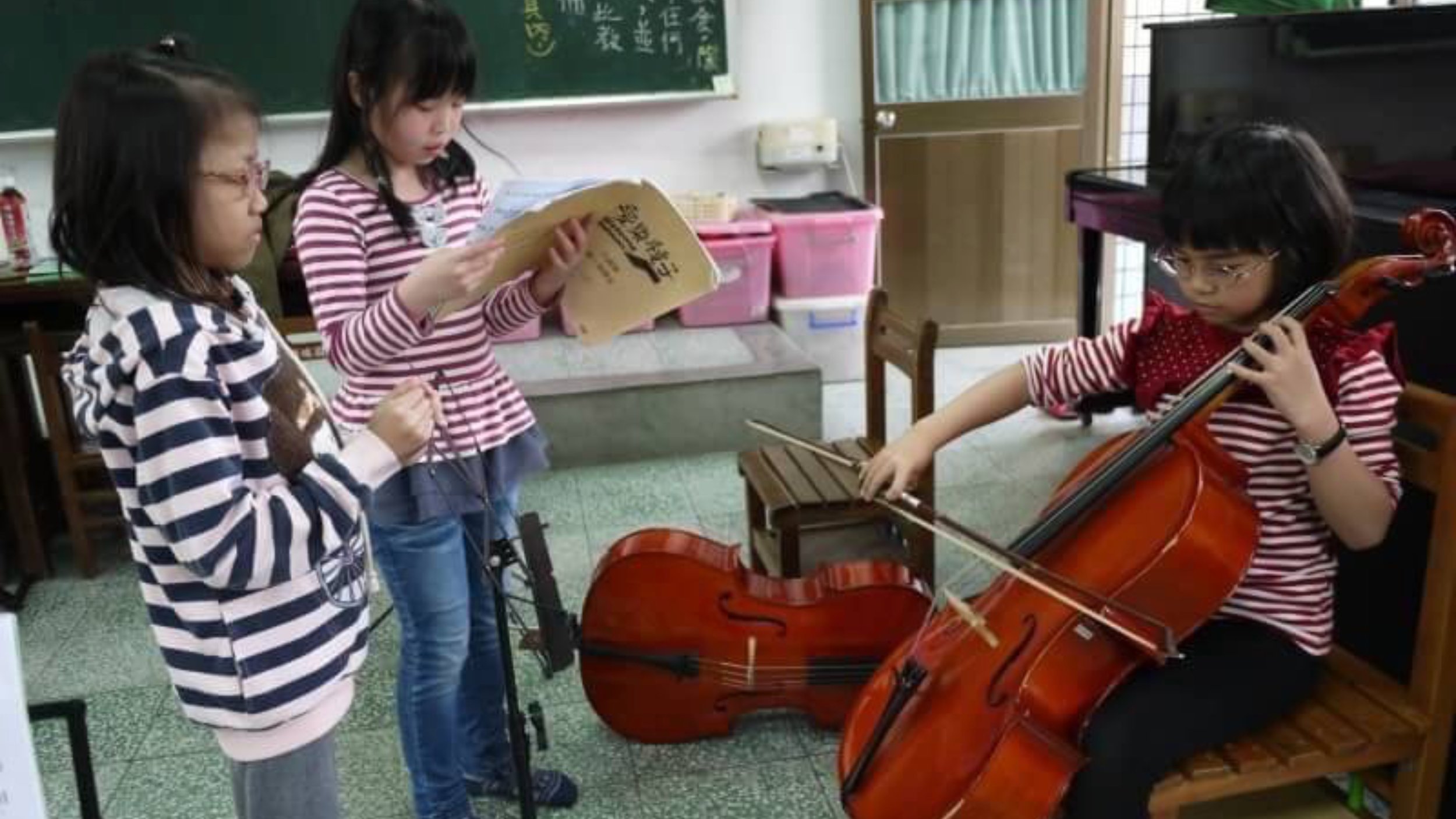 Three young girls practicing musical instruments in a classroom. One girl is playing the violin, and another girl is reading sheet music, while the third girl holds a music stand. The classroom has a blackboard and storage items in the background.