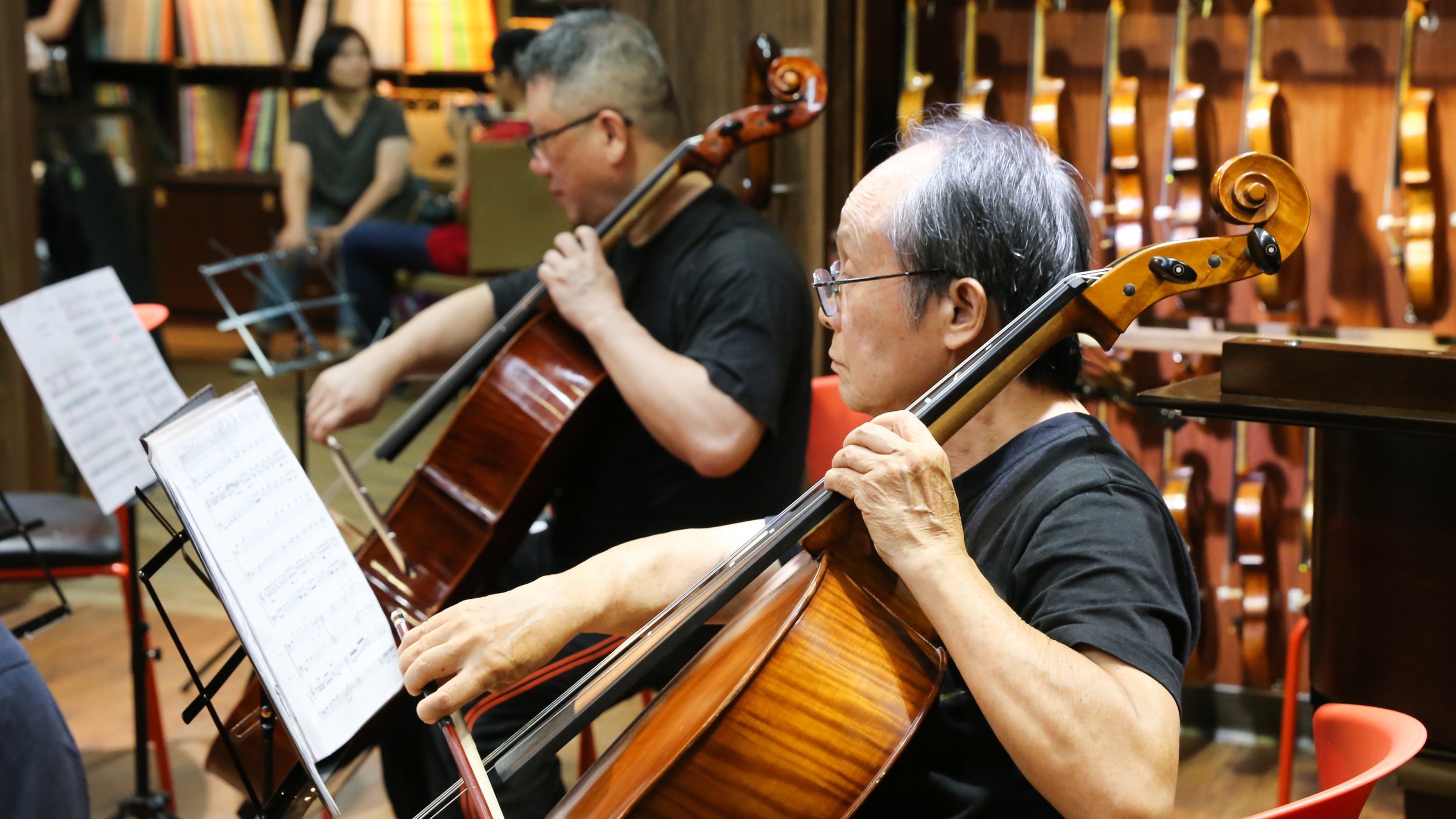Two elderly women and a man playing violoncellos during a music rehearsal in a room with wooden walls and shelves filled with books.