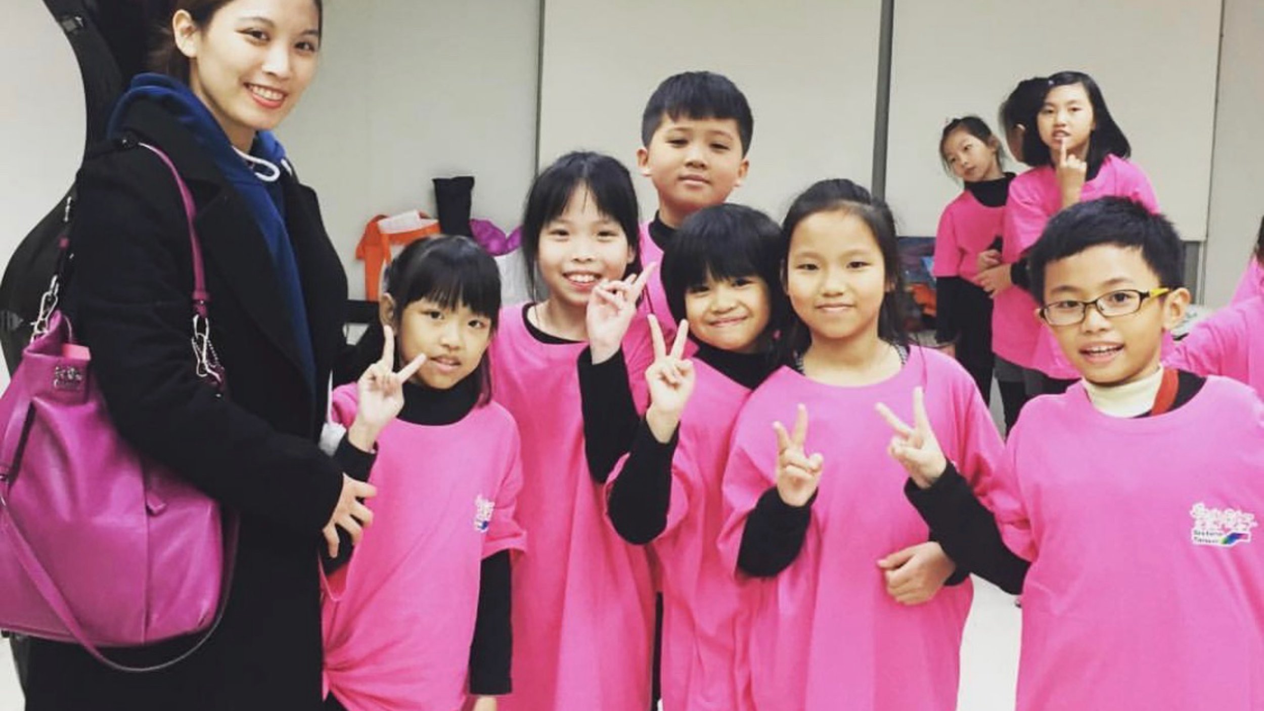 A group of children and a woman posing together with peace signs. The children are wearing pink T-shirts, and the woman is wearing a black jacket with a pink bag.