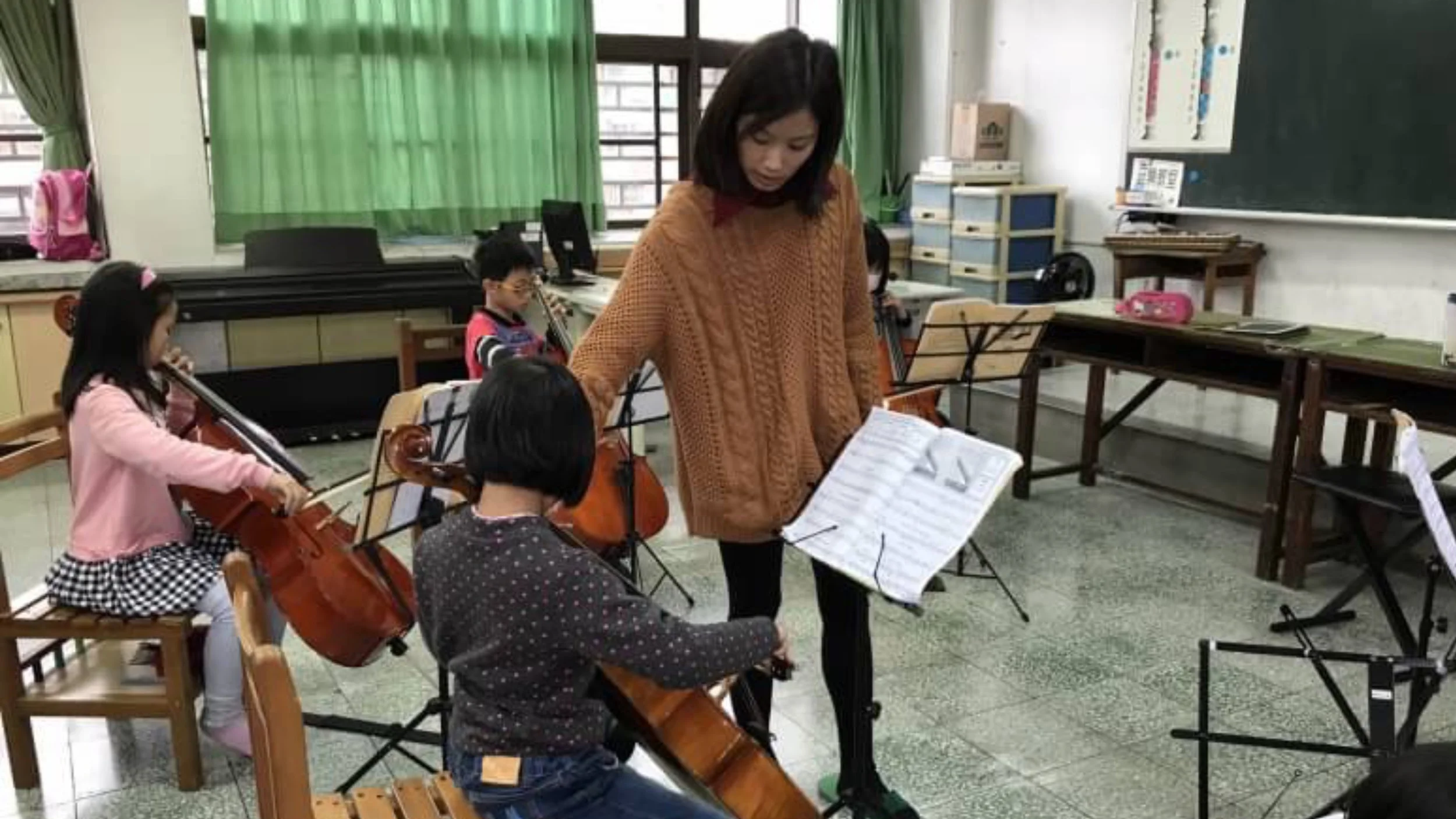 A music classroom with young children practicing string instruments, including violins and cellos. An adult instructor, possibly a teacher, looks at sheet music. The classroom has green curtains, desks, and educational posters.