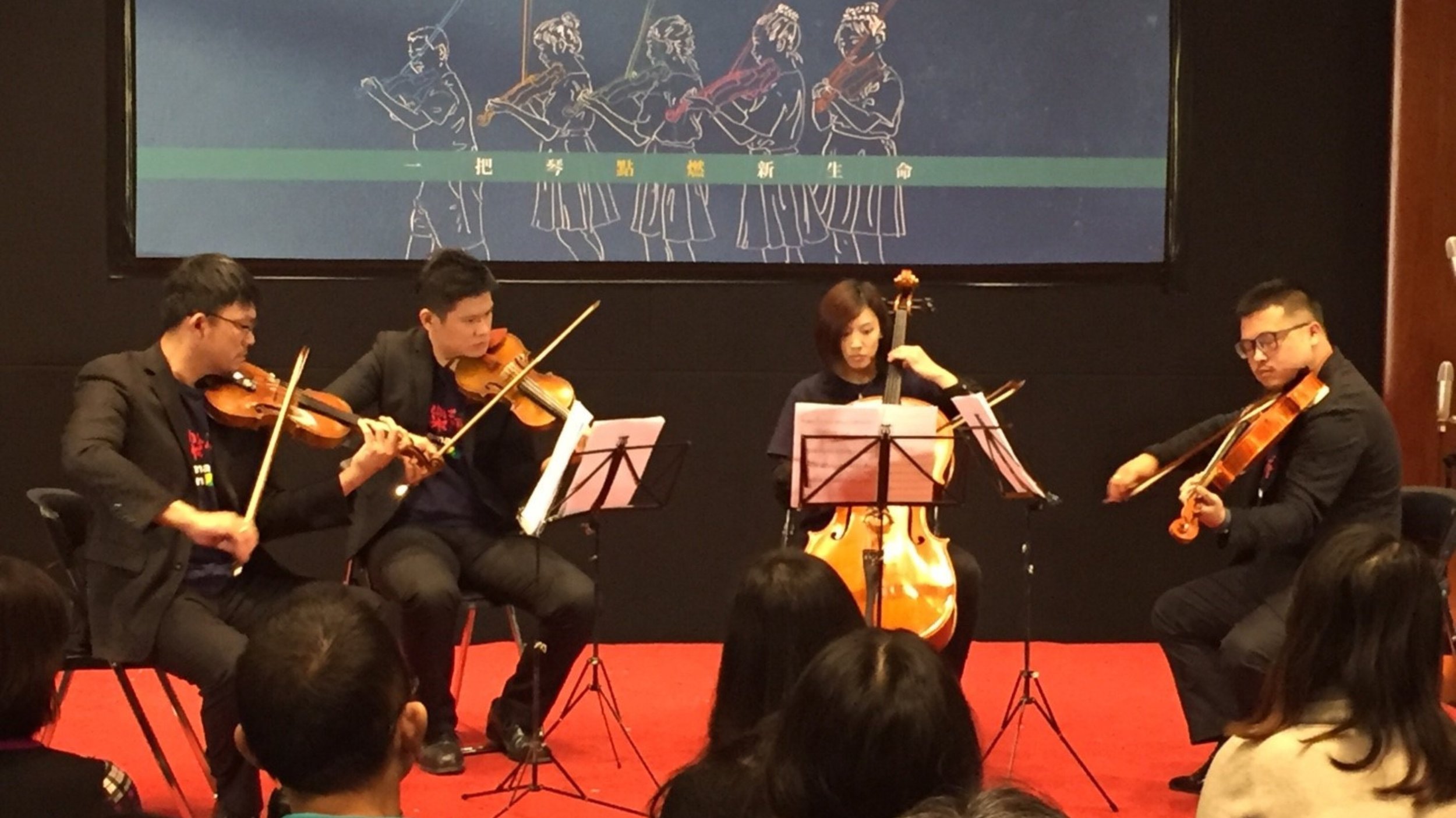 A group of four musicians playing string instruments on stage, with an audience watching. There is a large digital screen behind them displaying line art of children holding hands, with Chinese characters overlaying.