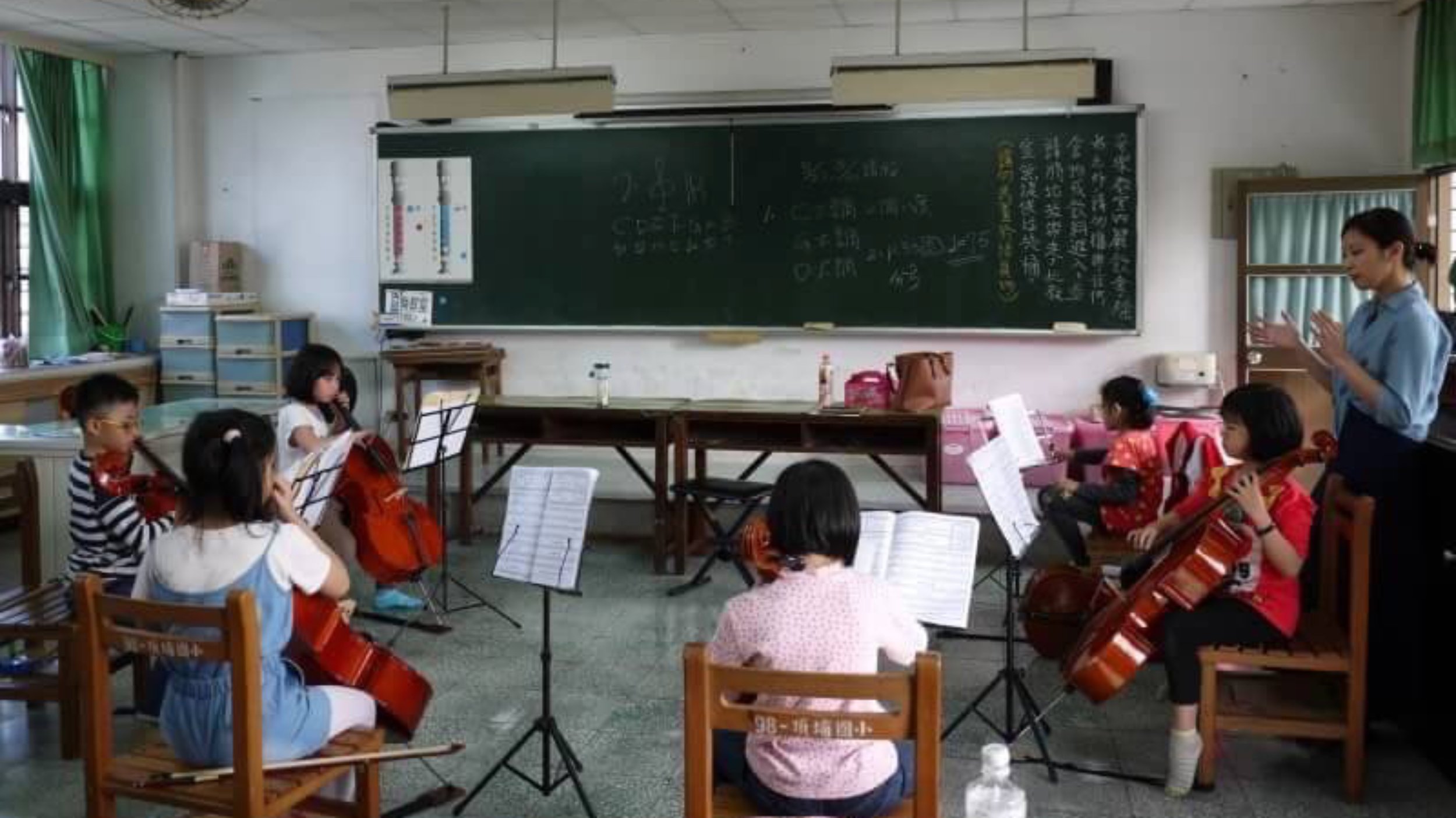 Young students practicing violins in a classroom with a teacher and a blackboard.