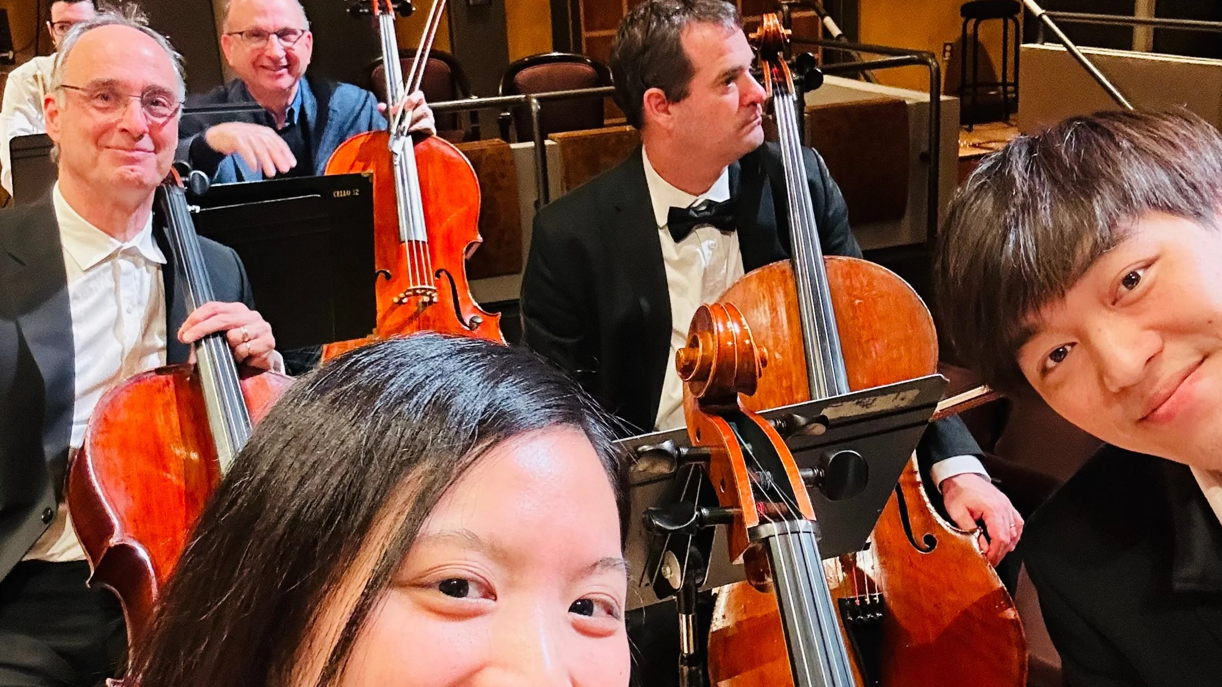 Photo of a group of musicians in formal attire, sitting with their string instruments, in an indoor setting, possibly backstage or at a concert hall.