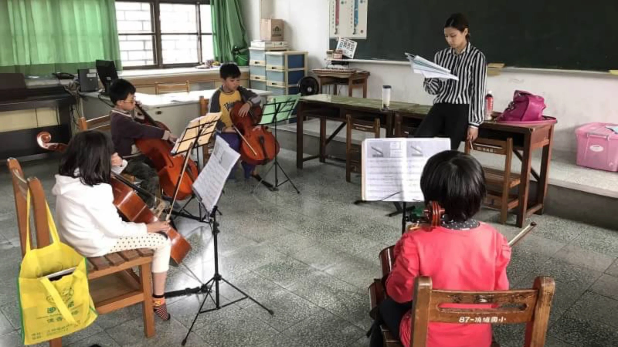 Students playing string instruments in a classroom with a teacher or instructor standing at the front.