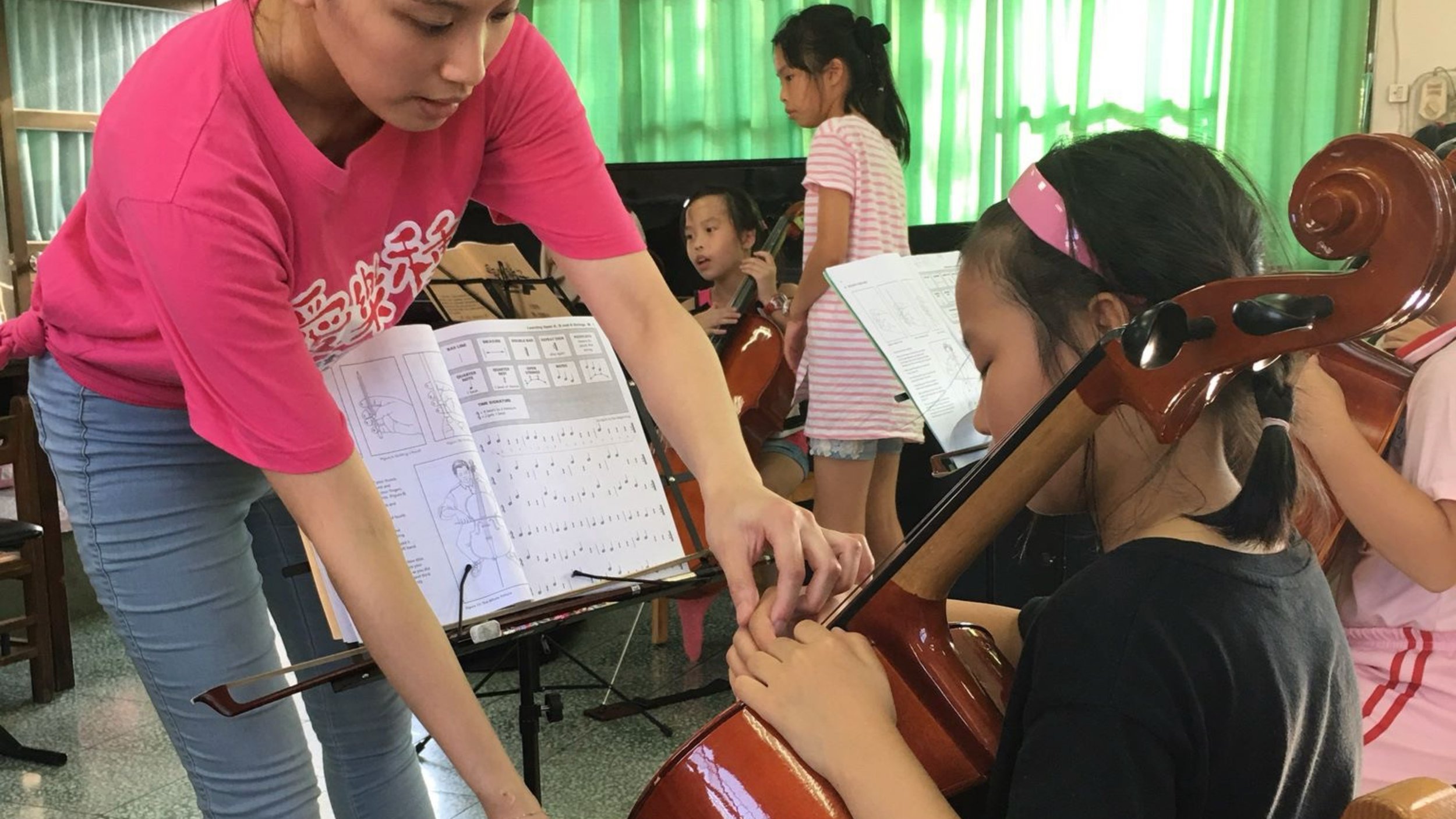 Music teacher instructs young girl playing the violin in a music classroom with other children practicing instruments in the background.