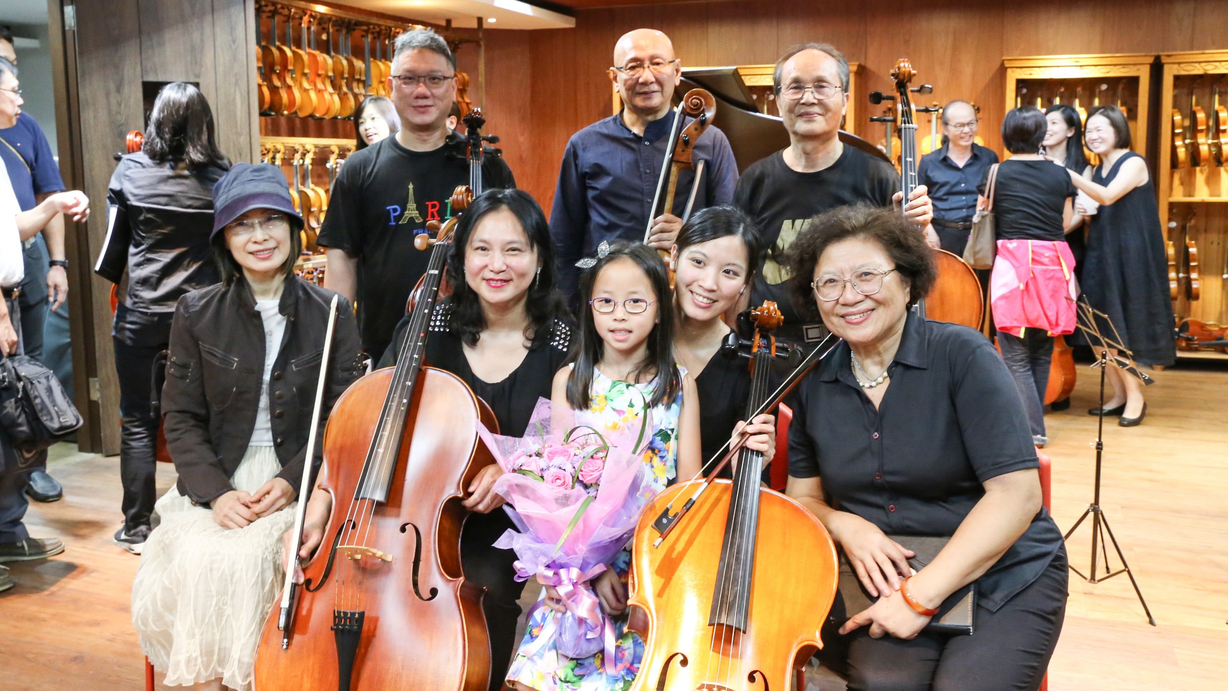 A group of musicians and children smiling with musical instruments, including cellos and violins, in a room with wooden floors and walls, with more musicians and violin shelves in the background.