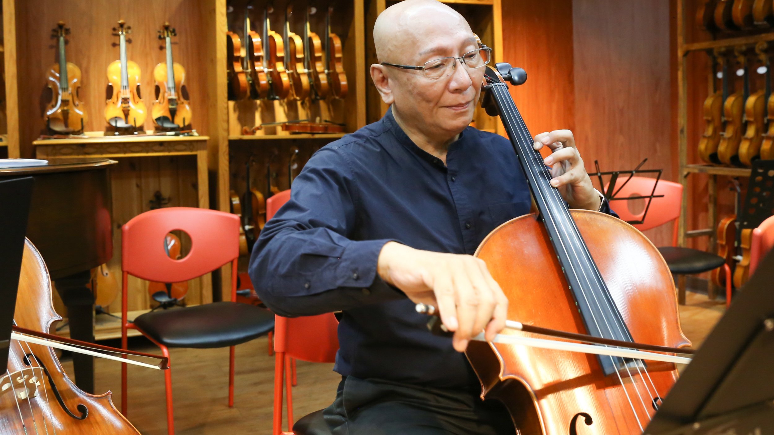 An elderly man with glasses playing a cello in a room filled with multiple violins hanging on wooden shelves.