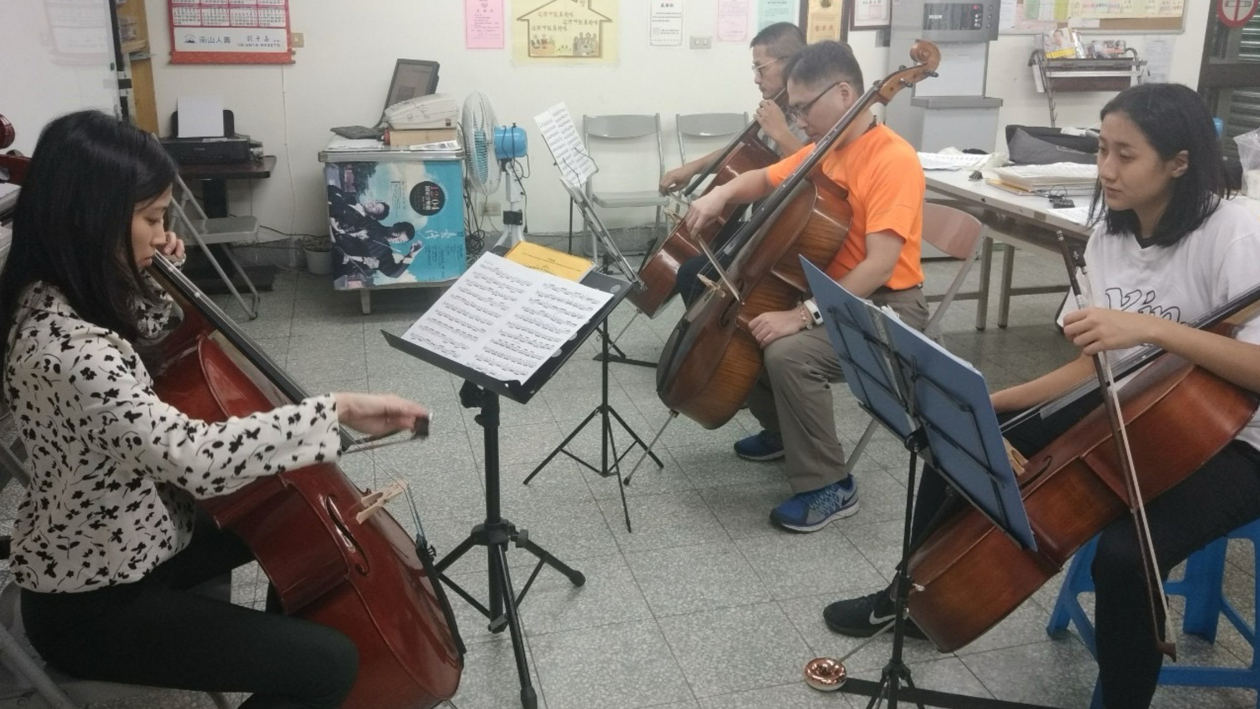 A group of four musicians, three men and one woman, playing classical string instruments including a cello and a violin, in a room with sheet music on stands, posters on the wall, and various office supplies in the background.