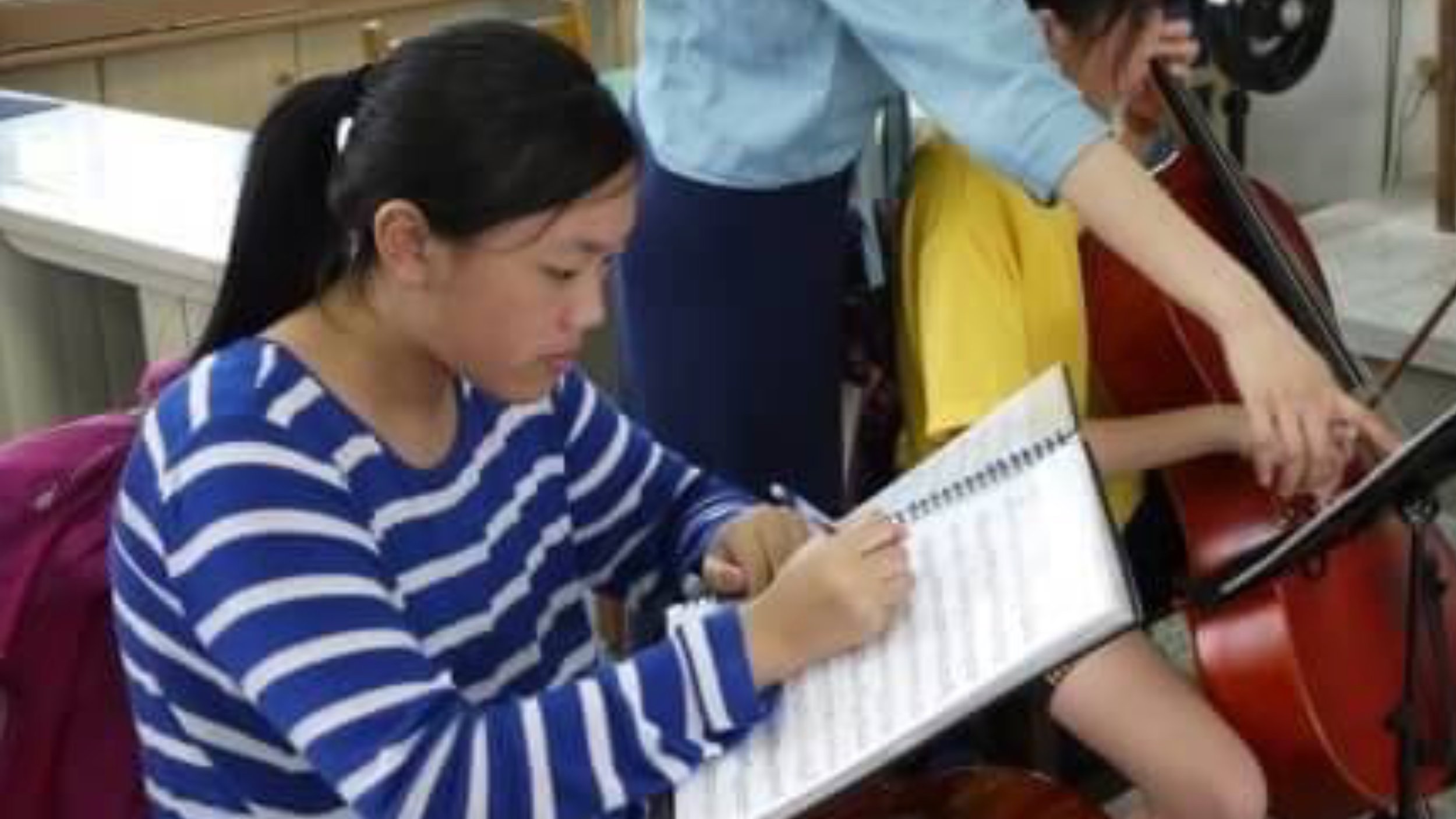 Two students sitting at a desk in a classroom, focused on writing and using a laptop.