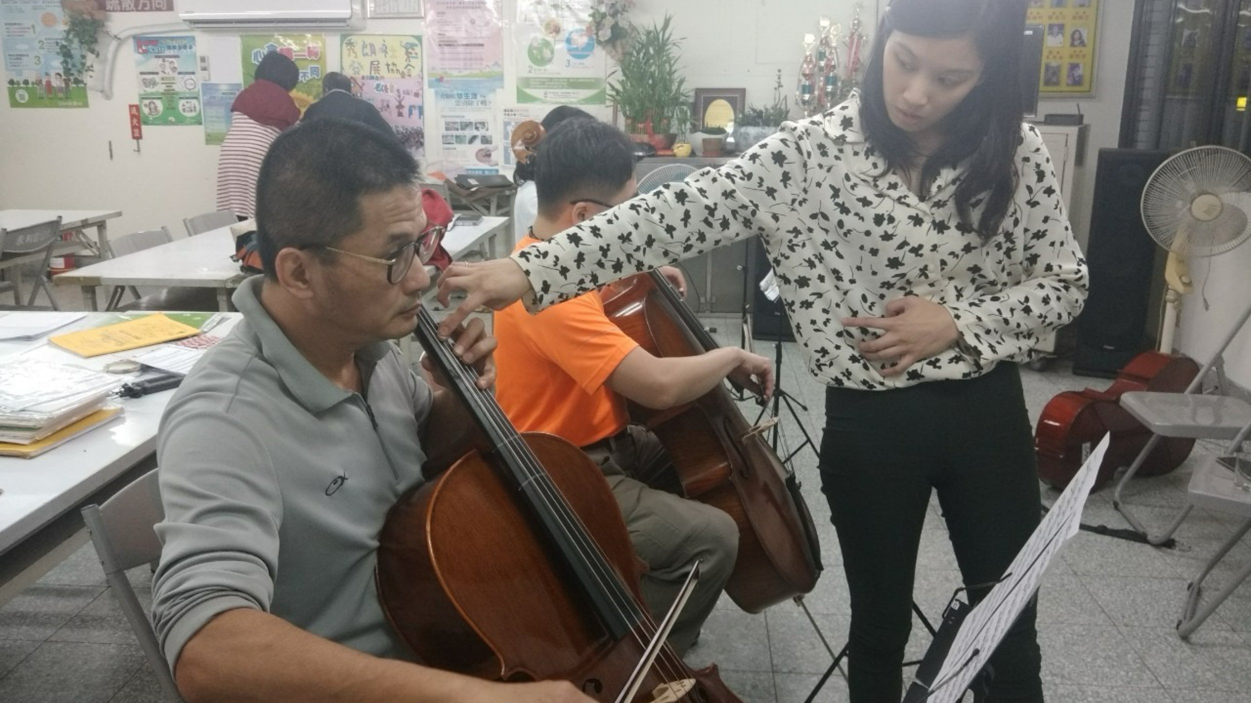 Woman instructing a man playing a cello in a classroom with other seated musicians, posters on the wall, and musical instruments nearby.