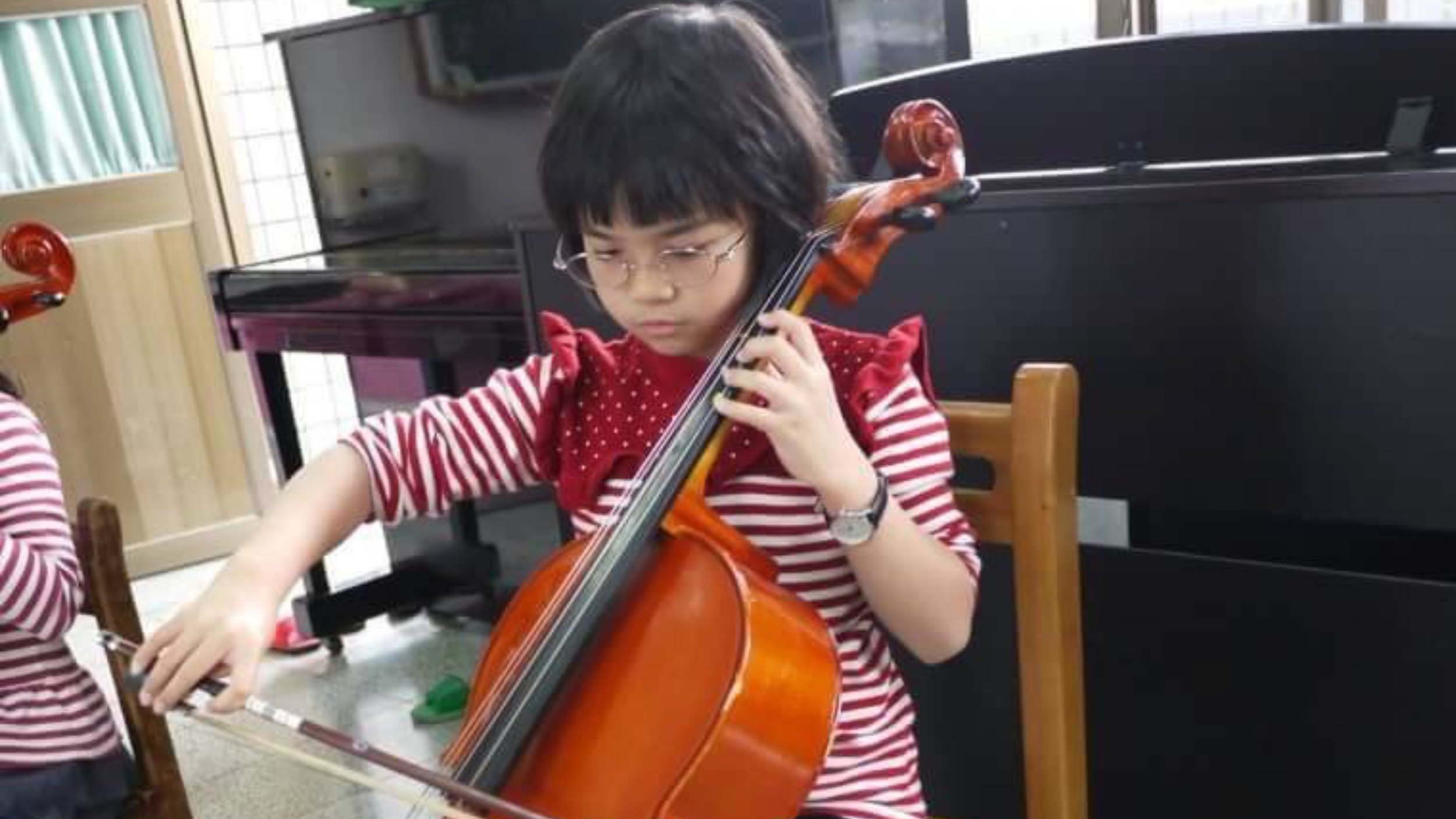 A young girl with short dark hair and glasses playing a cello indoors, wearing a red and white striped shirt and a red polka-dot scarf.