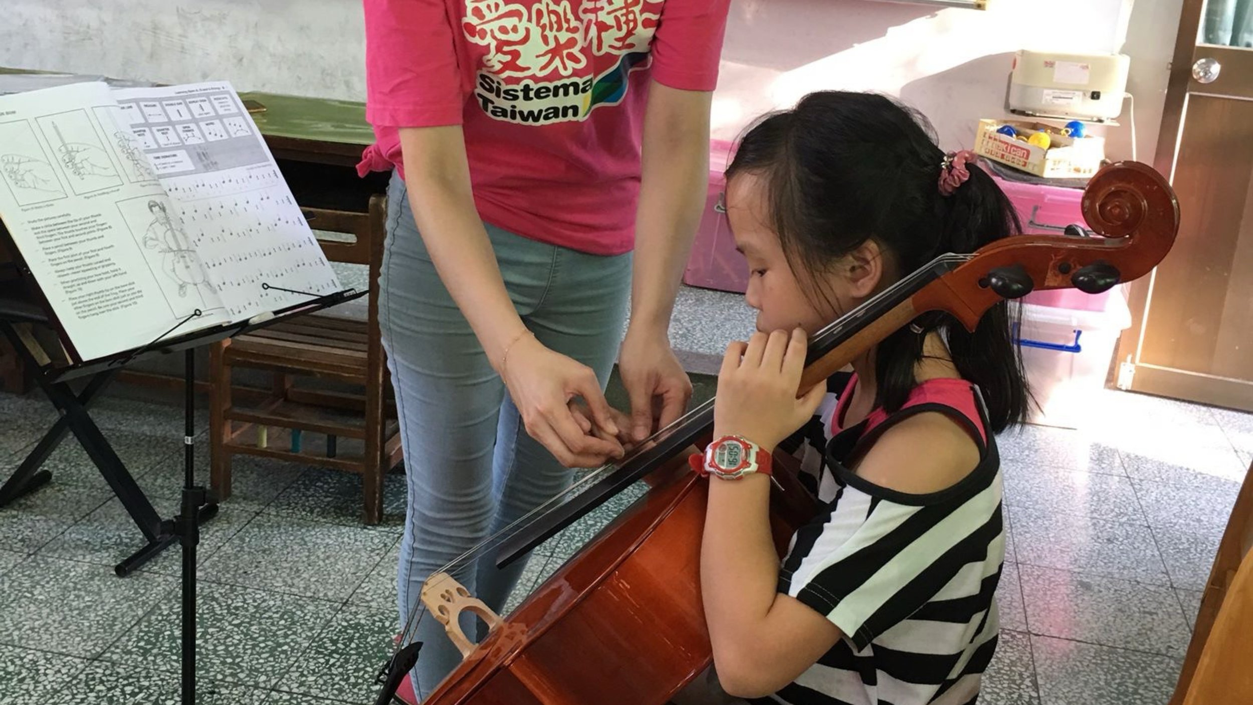 A young girl playing the violin while an adult helps her with her fingering, in a room with a music stand and sheet music.