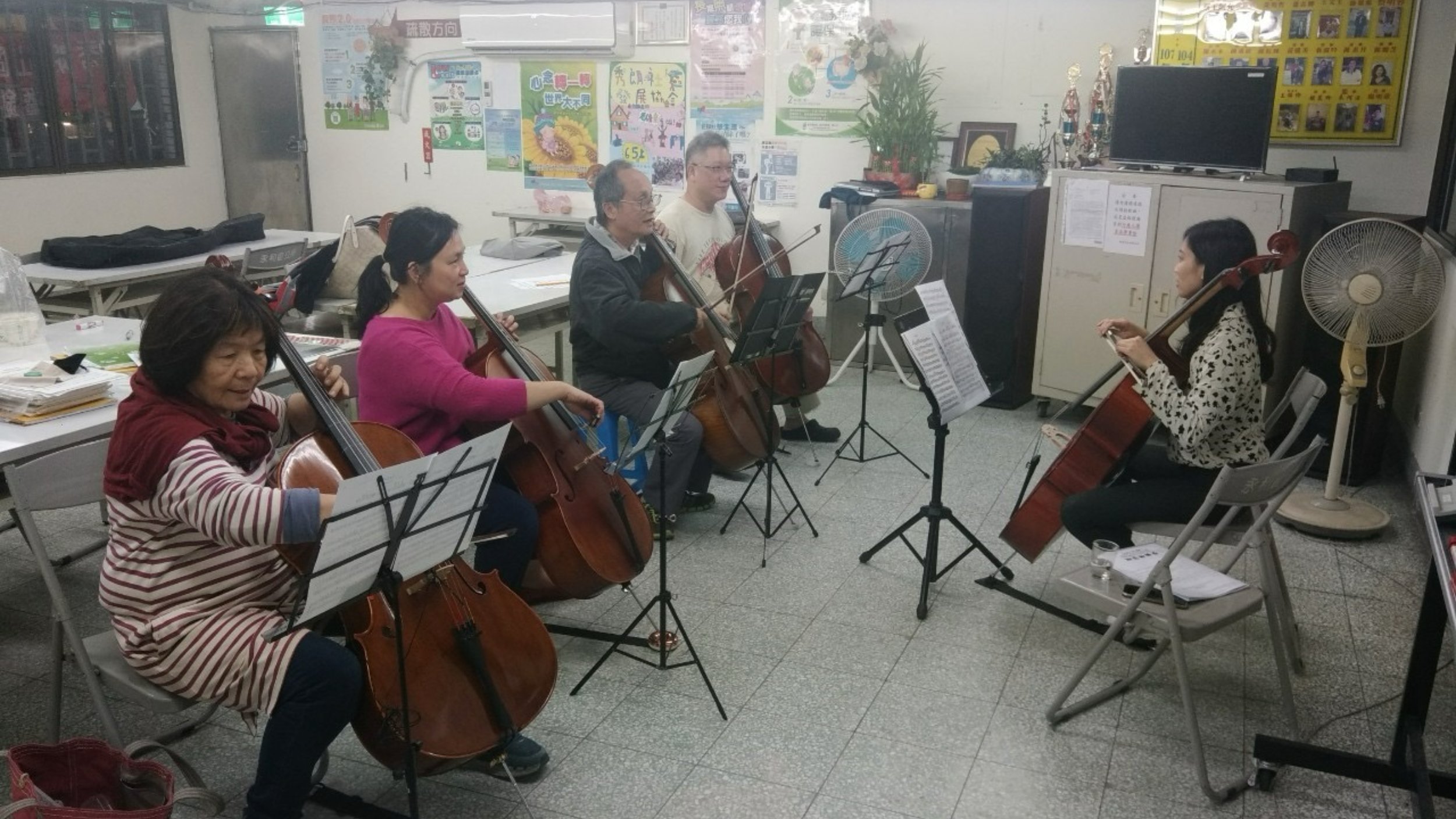 A group of five people playing string instruments, including violins and cellos, in a room with posters on the wall, a television, and a fan, indicating a practice or rehearsal session.